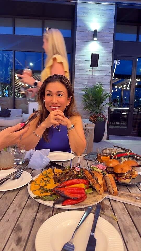 Smiling woman in a blue top sits at an outdoor restaurant table with a large platter of grilled meat, peppers, and buns; another guest holds a phone nearby.