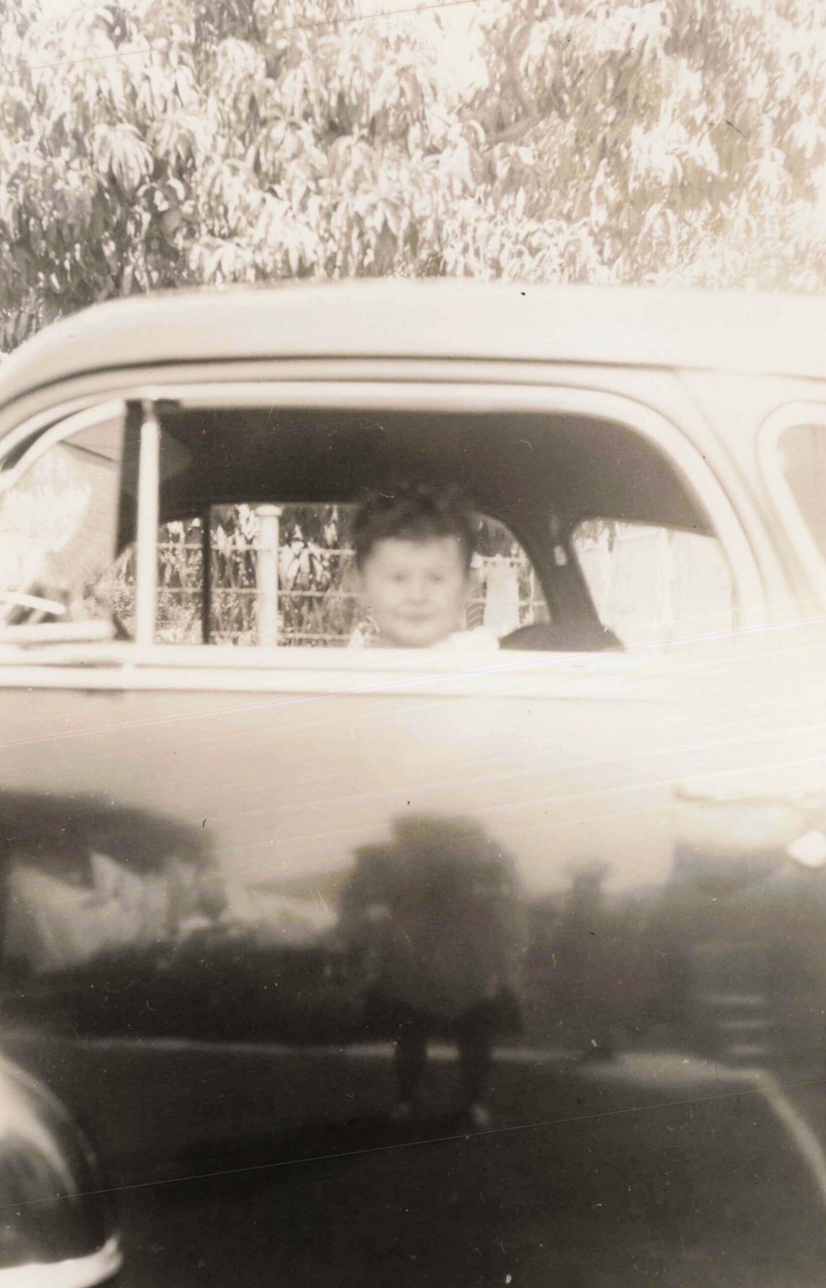 Person sitting in the driver's seat of a vintage car, window down, with trees in the background (old photograph).