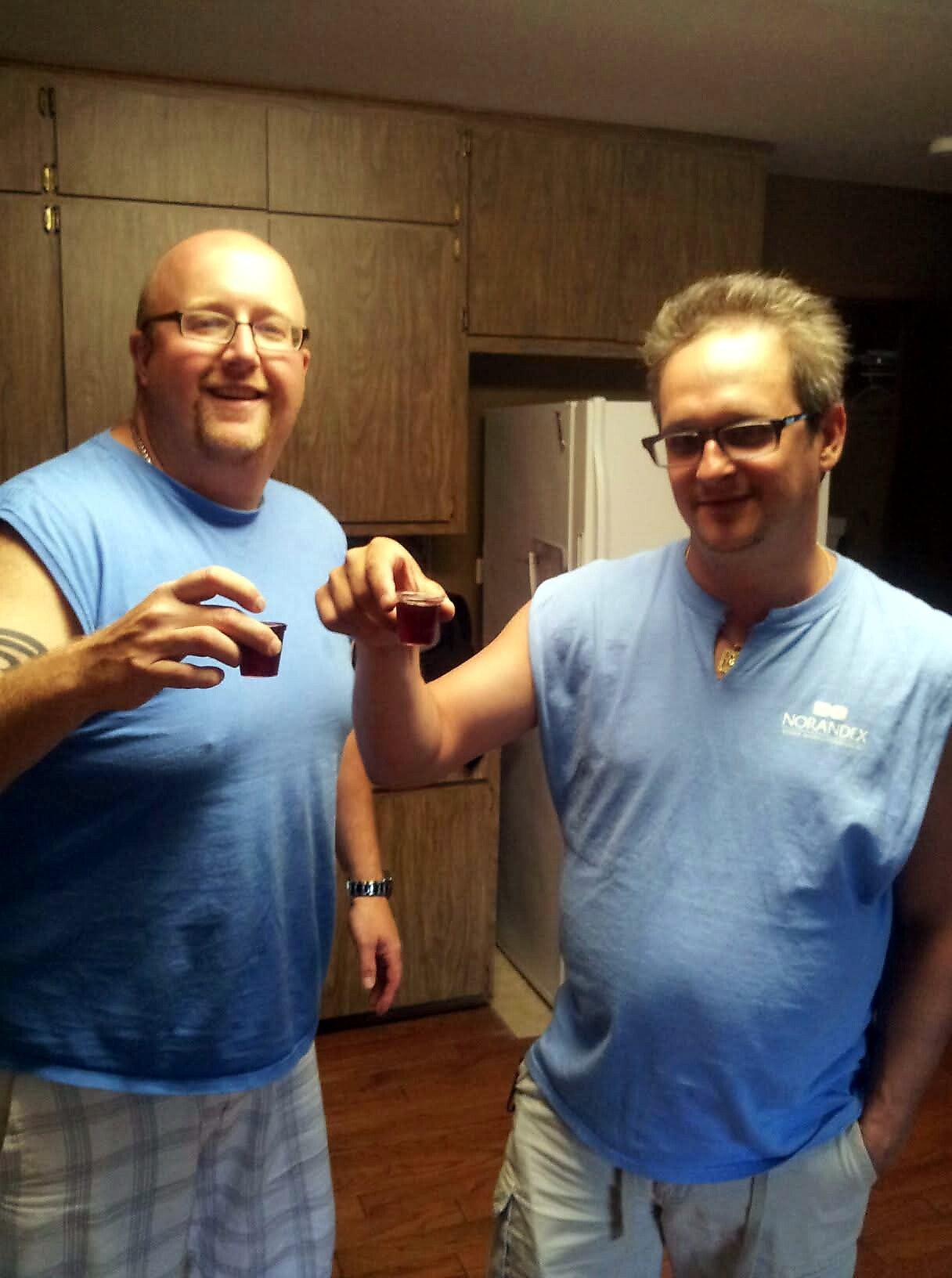 Two men in blue T-shirts toast with small red shot glasses in a kitchen setting, smiling at the camera.