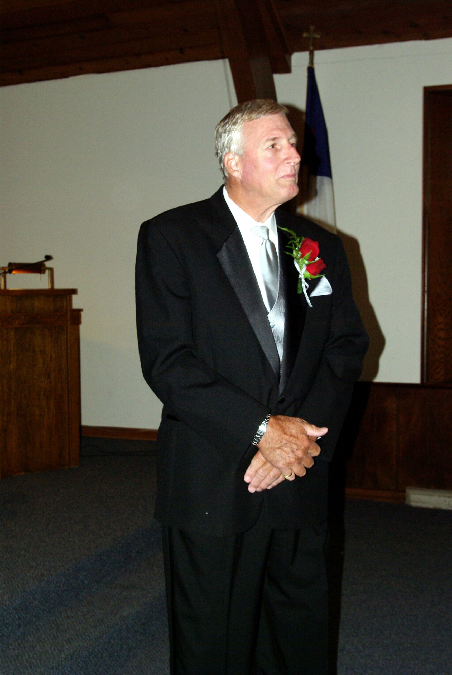 Older man in a black tuxedo with a red rose boutonniere standing in a hall beside a podium.