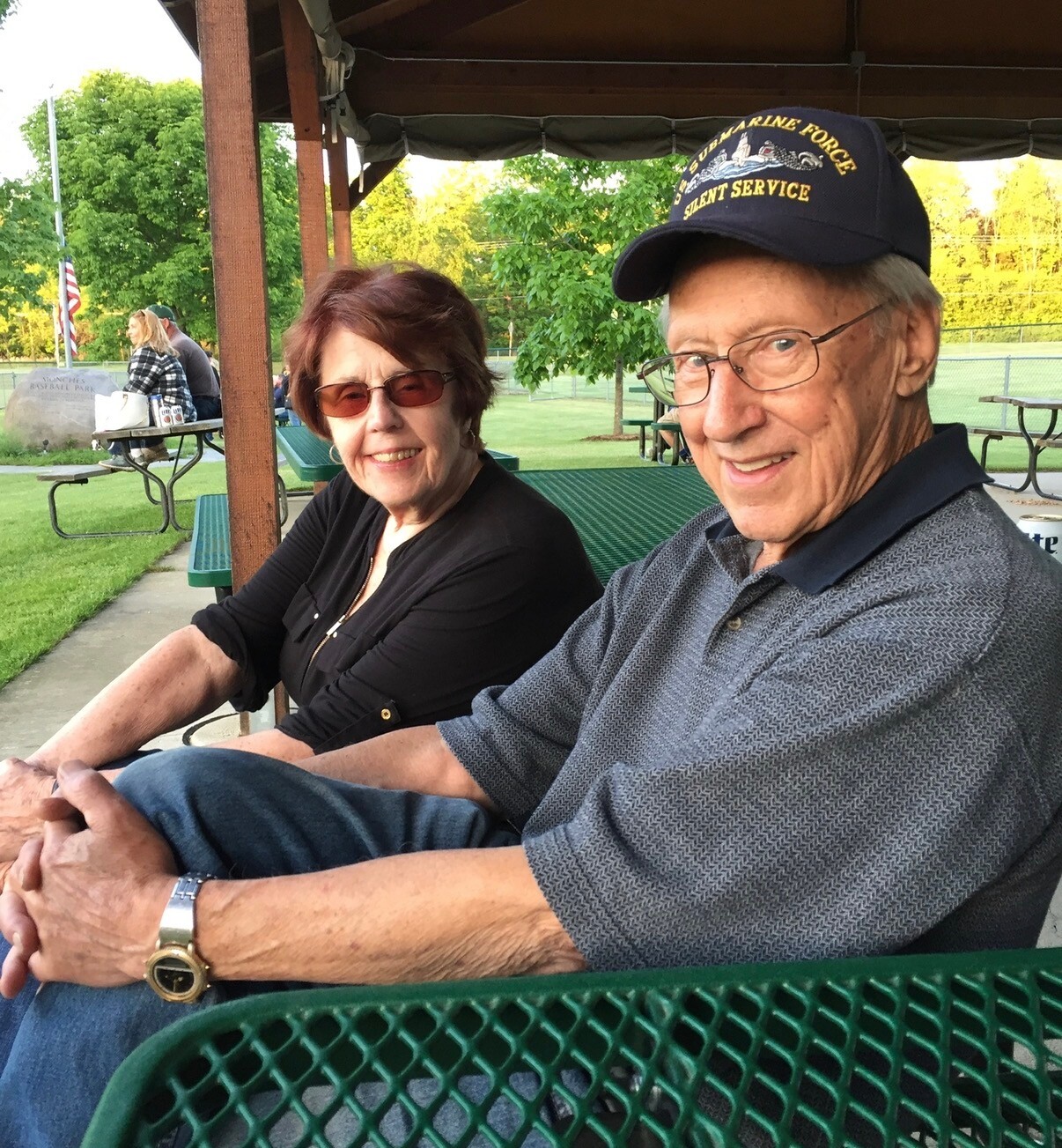Older couple seated at a park pavilion, holding hands and smiling at the camera, with trees and a field in the background.