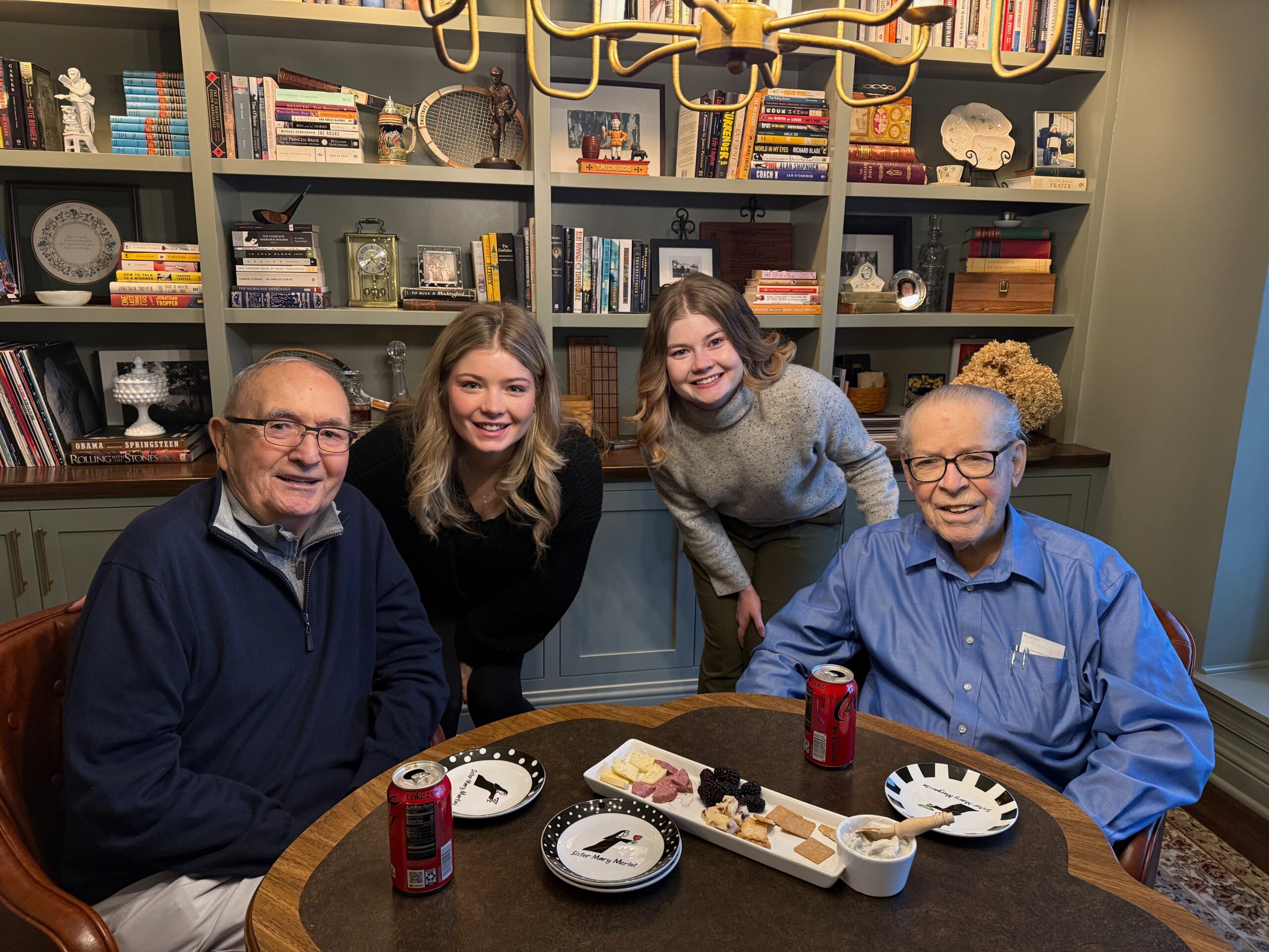 Group of four people sitting around a round table with a cheese platter and sodas, in a home library filled with books and decor behind them.