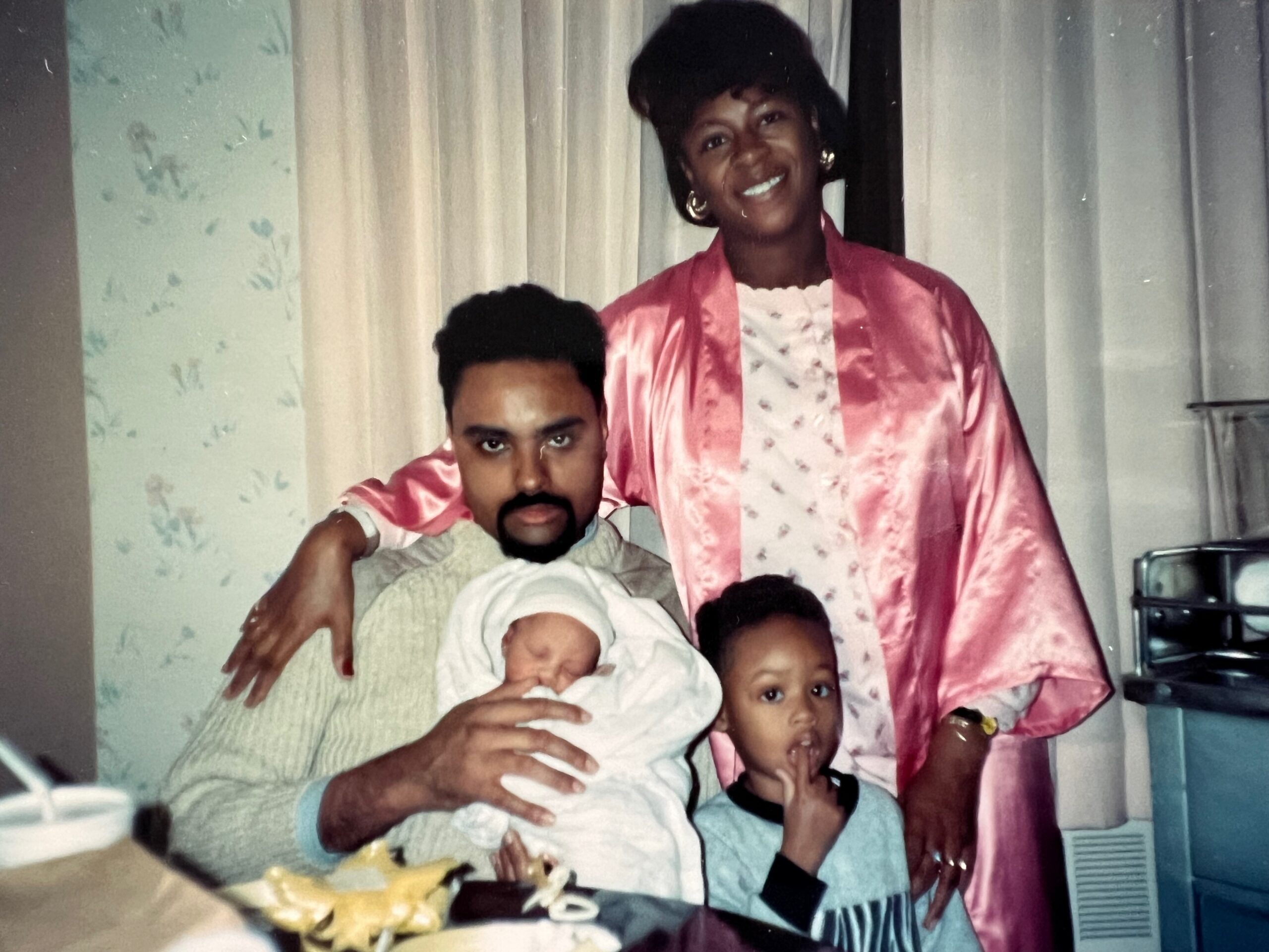 Smiling woman in a pink satin robe stands behind a man holding a wrapped baby, with two children nearby and a cake on the table in a home setting.