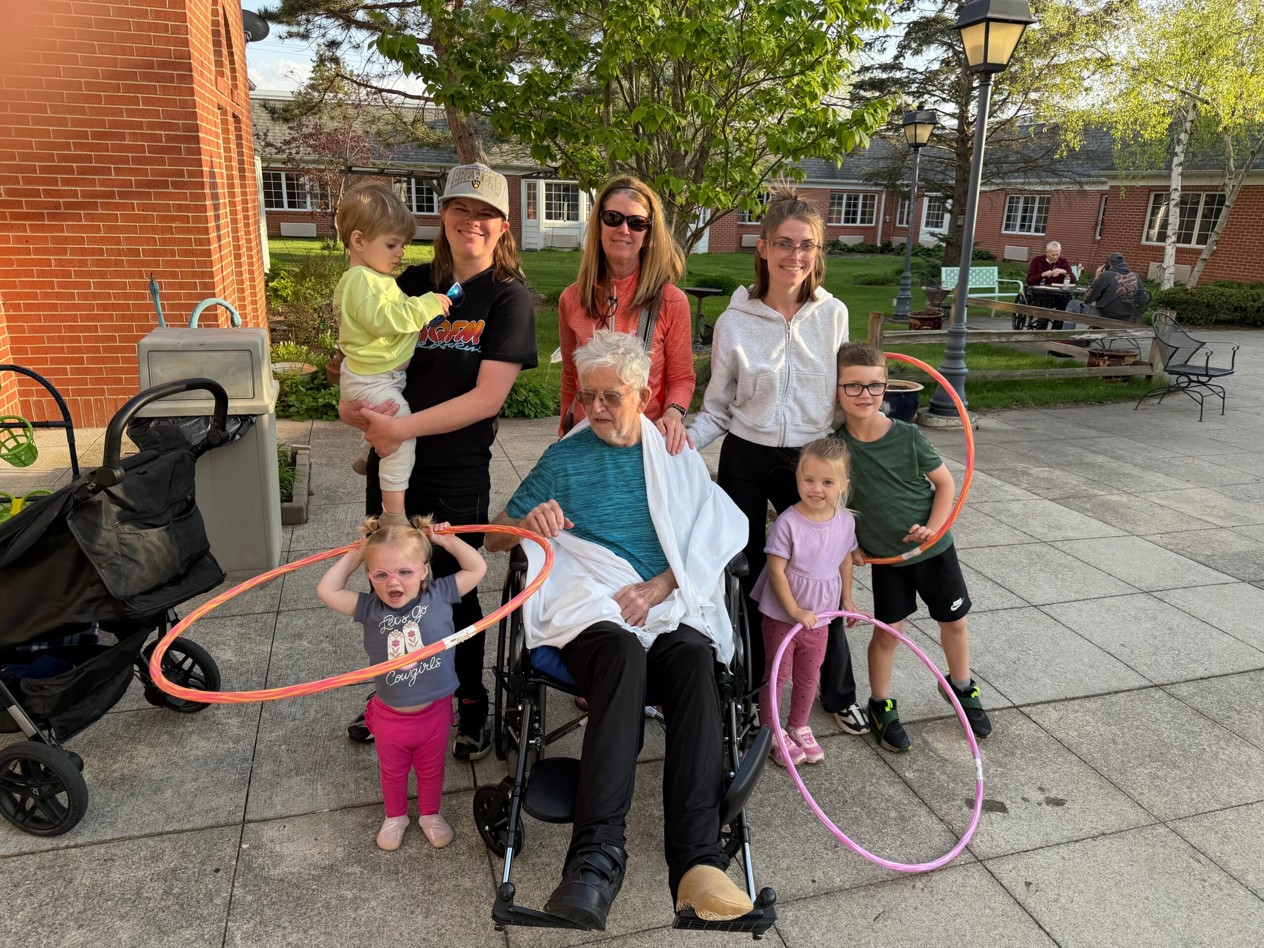 Group photo outdoors in a courtyard: adults and children gathered around an elderly man in a wheelchair, with colorful hula hoops nearby.
