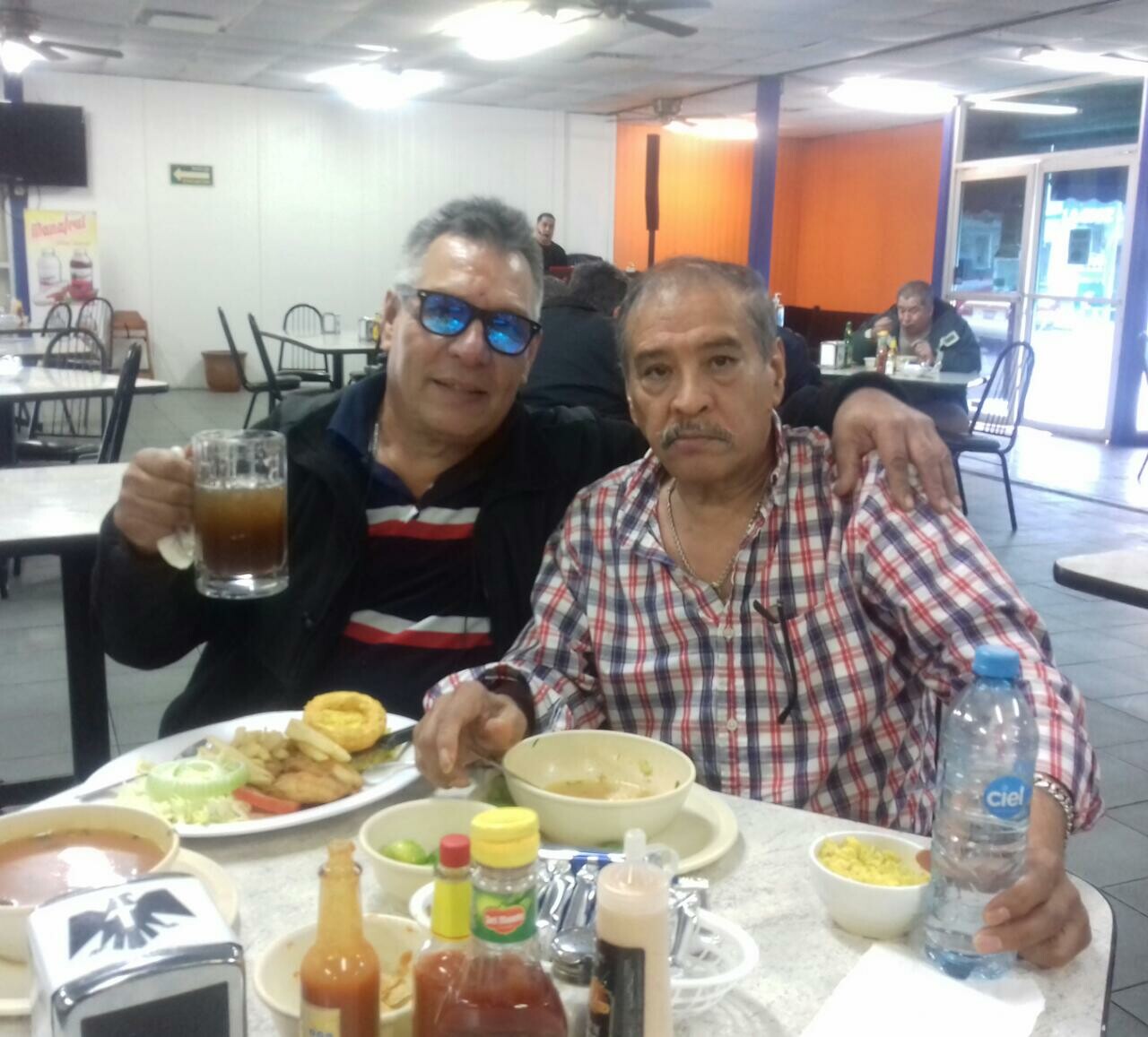 Two middle-aged men sit at a casual restaurant table, one holding a beer mug and wearing sunglasses, the other in a plaid shirt looking at the camera.
