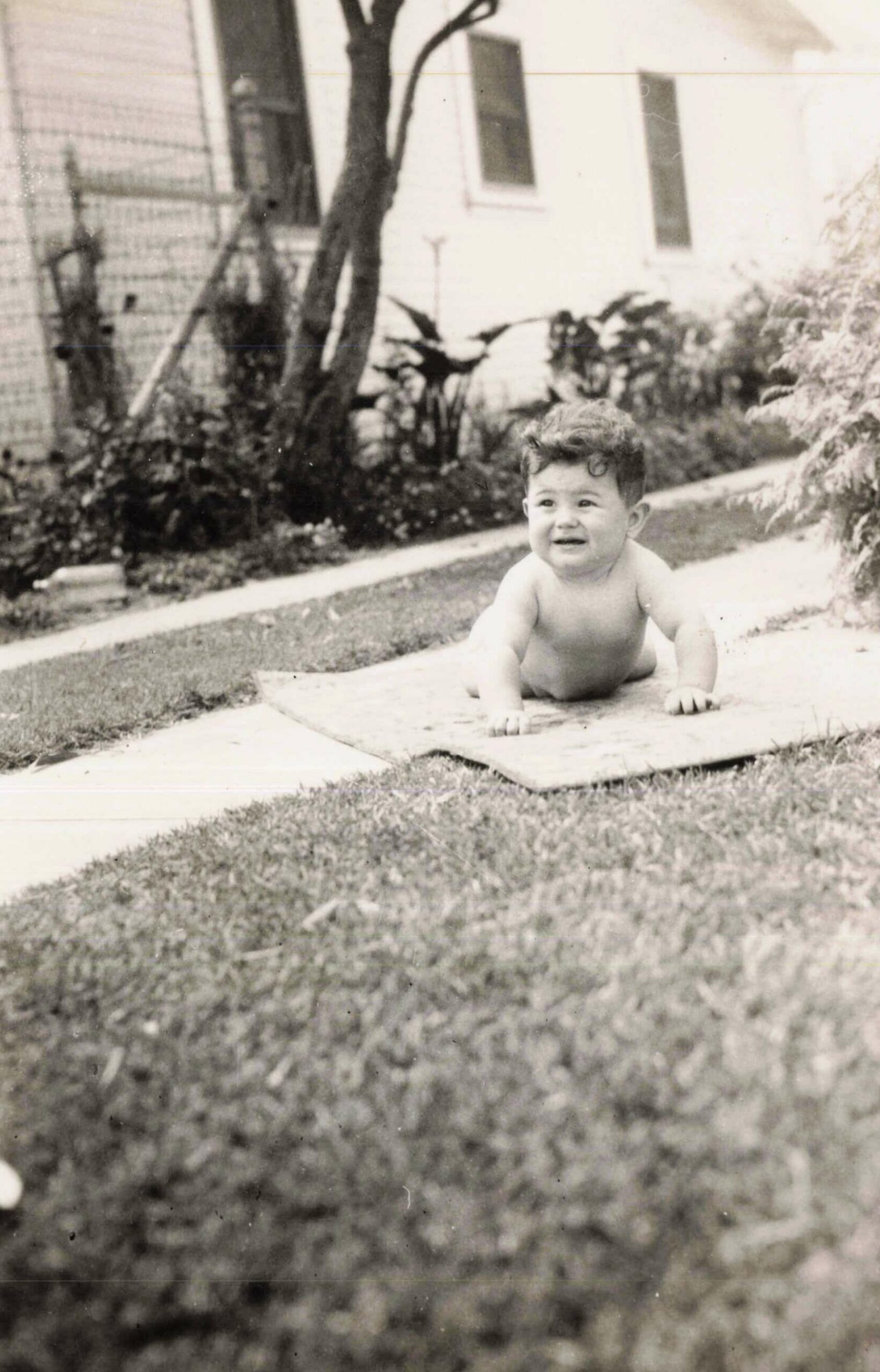 Smiling baby crawling on a stone path in a backyard, with a white house and plants in the background.