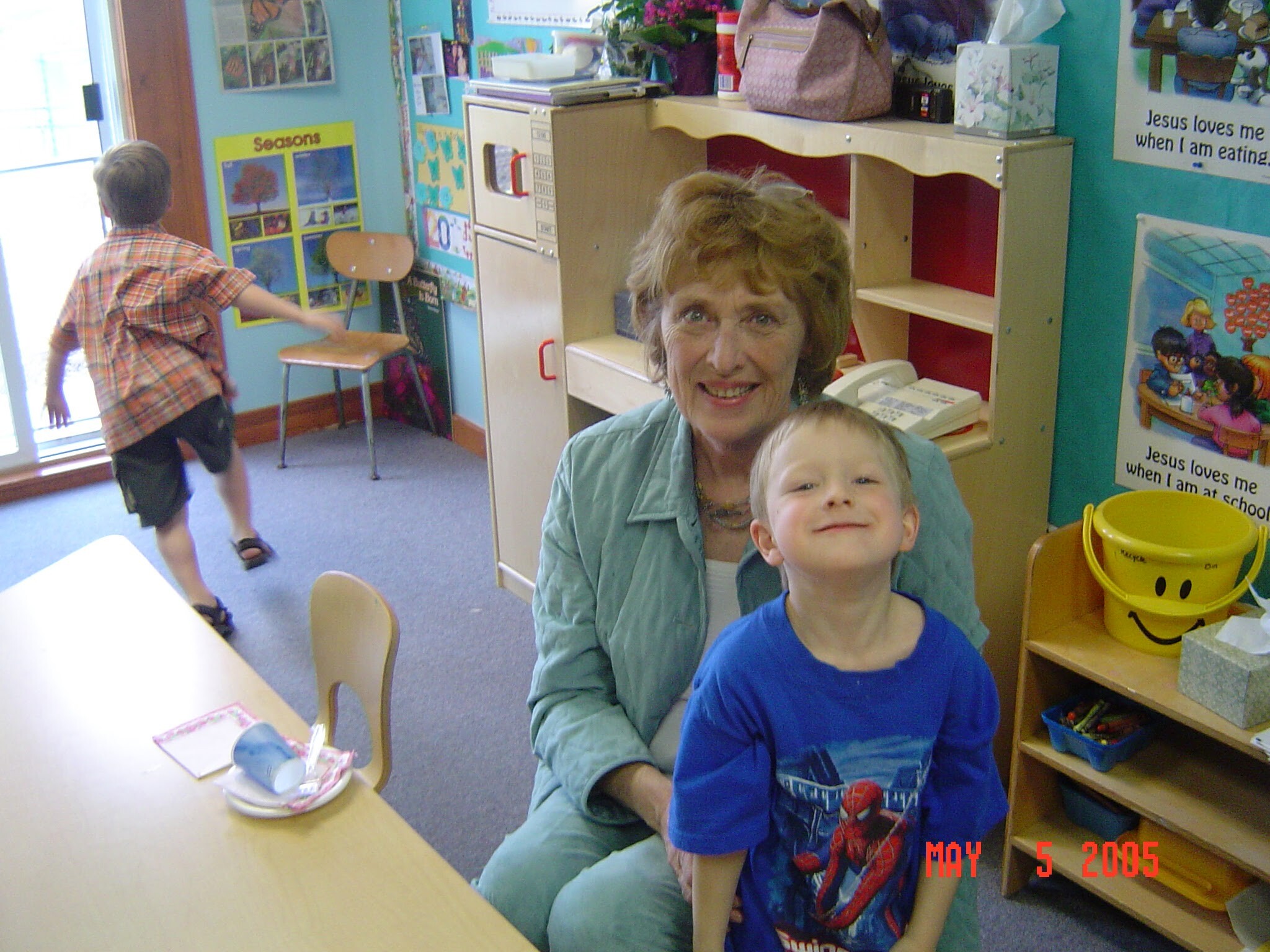 Smiling woman beside a child in a colorful classroom; a boy runs toward the left doorway, posters and shelves visible.