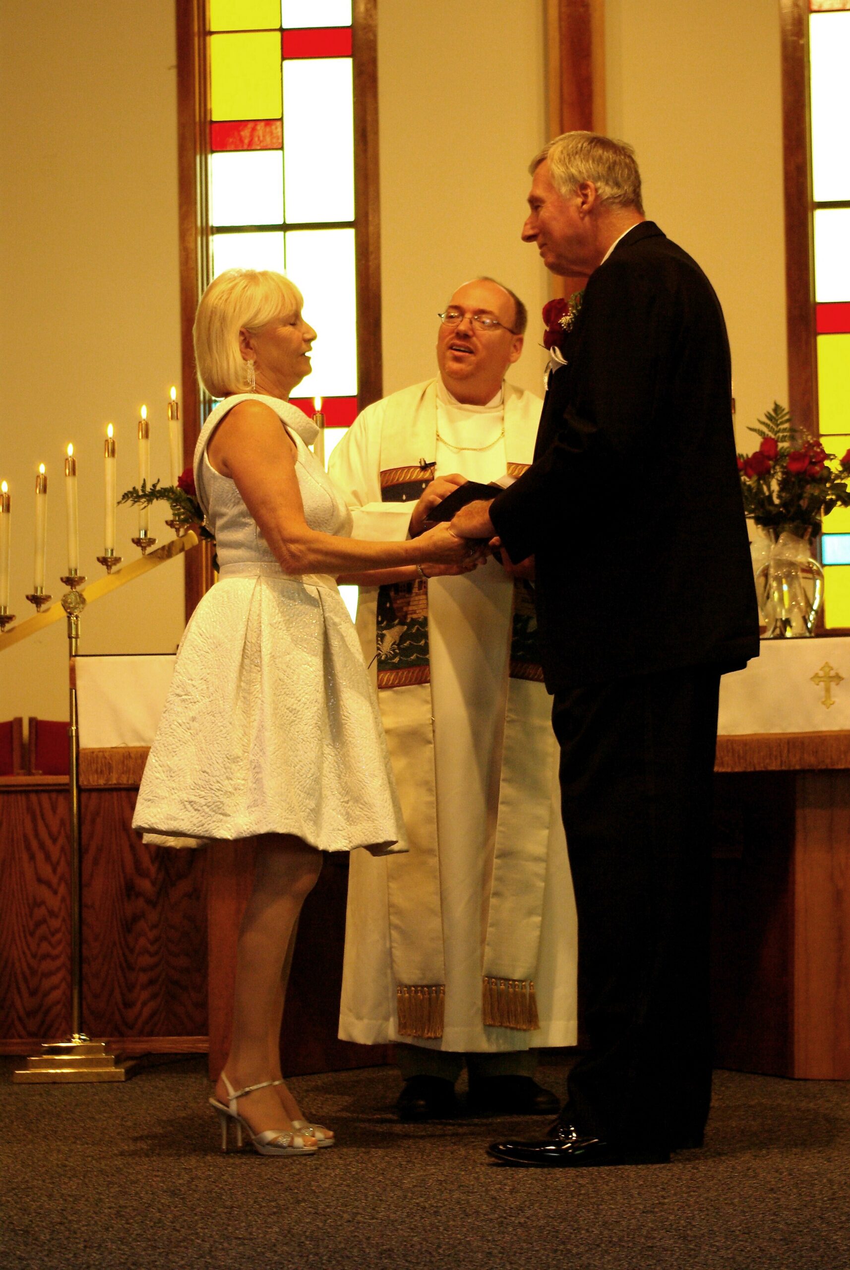Bride in a light blue dress and heels exchanges rings with a groom in a black suit during a church wedding, officiant standing between them with stained-glass windows and candles in the background