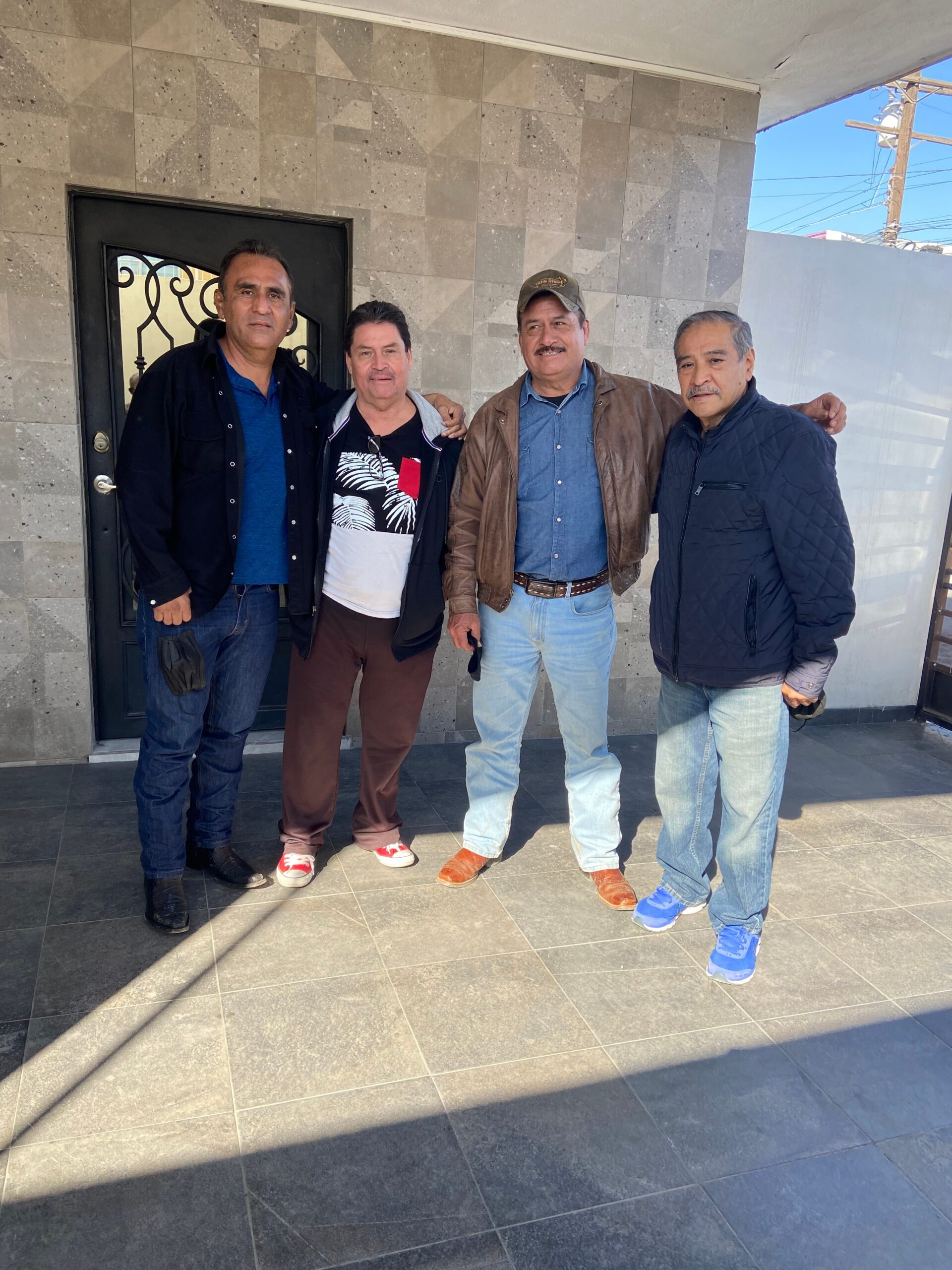 Four men stand together outside a tiled wall and doorway, posing for the photo with arms around each other.