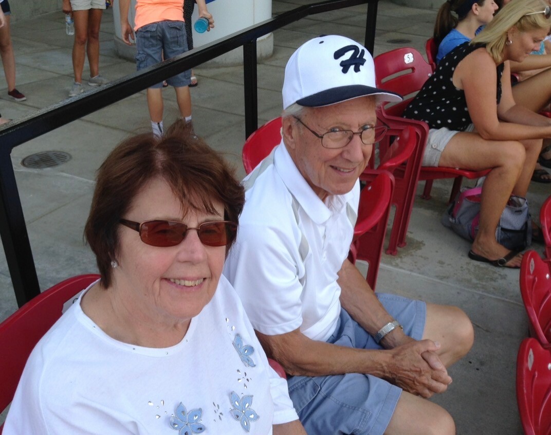 Smiling older couple sitting in red stadium seats at an outdoor event; woman in a white floral shirt, man in a white polo and cap