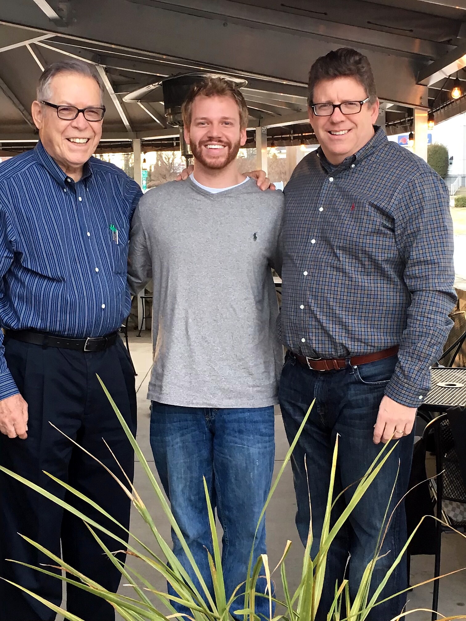 Three men smiling and posing together under a covered area, with plant fronds in the foreground.
