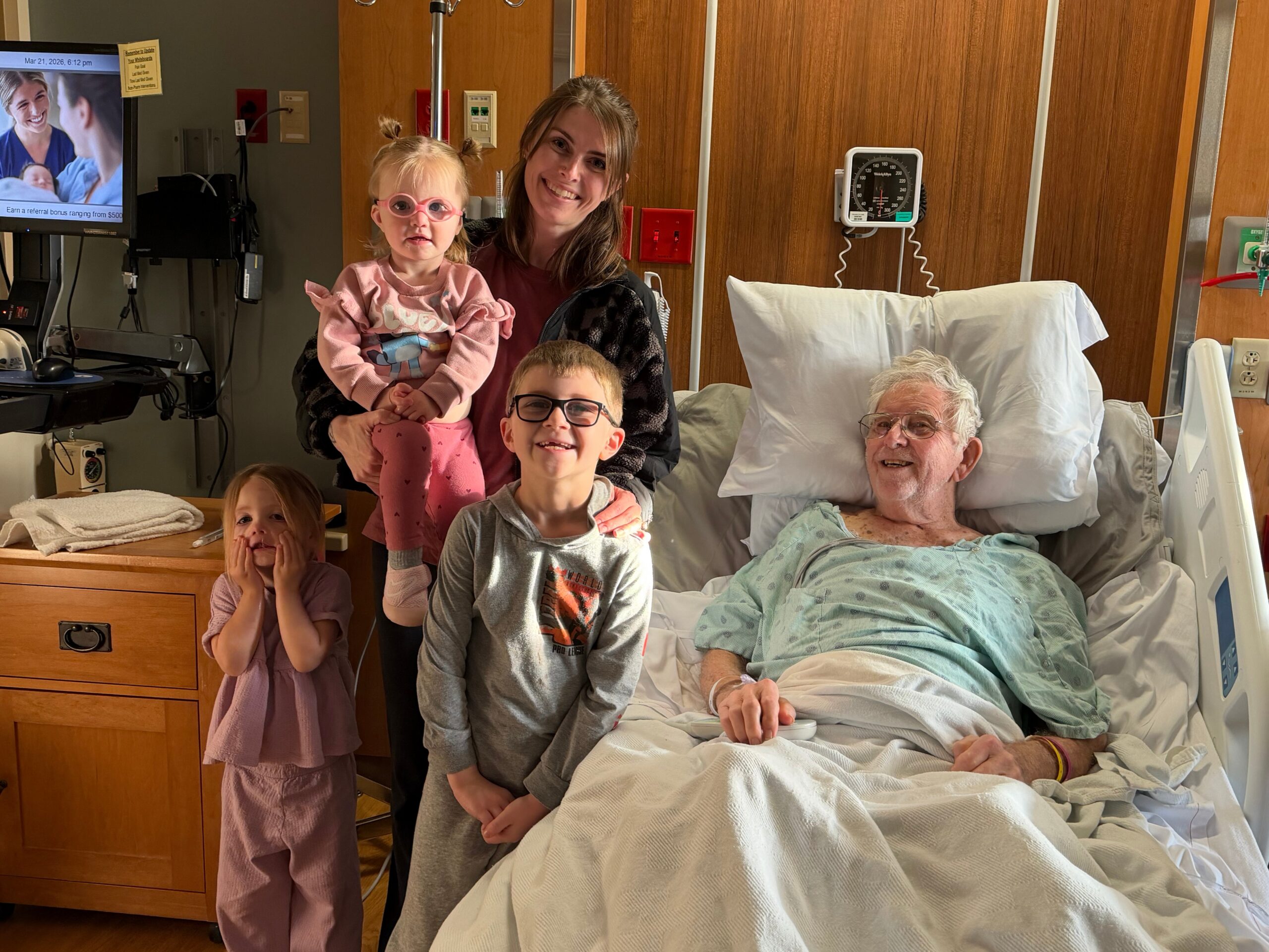 Family in a hospital room posing for a photo: an elderly man in bed with a blanket, a smiling woman holds a small pink-clad girl, and a boy stands in front with glasses nearby.