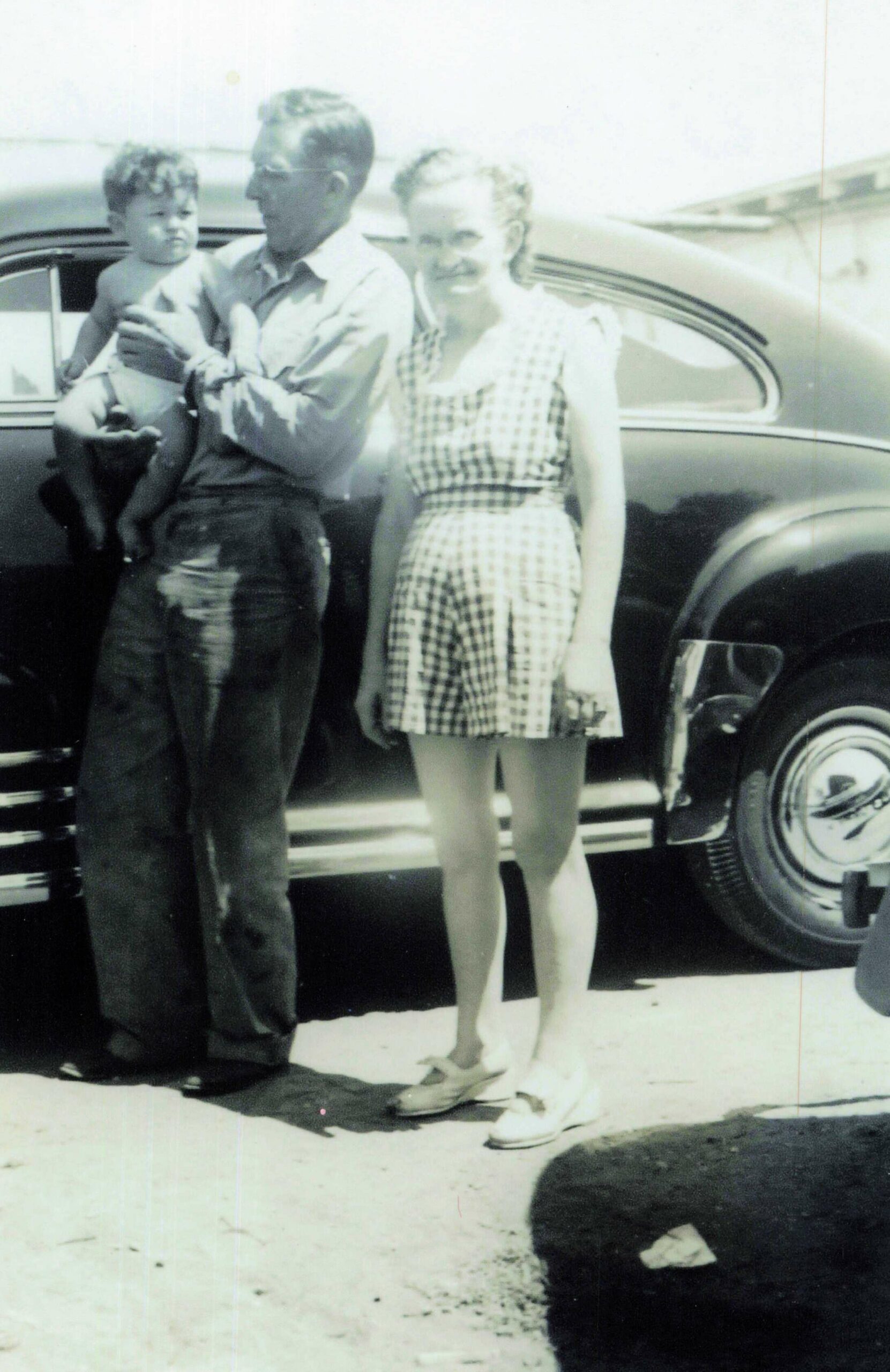Man holding a toddler on his hip while a woman stands beside them in front of a classic car; appears to be a family photo from mid-20th century.