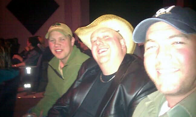 Three men smiling at the camera in a dimly lit bar; the center man wears a straw cowboy hat and a black jacket.