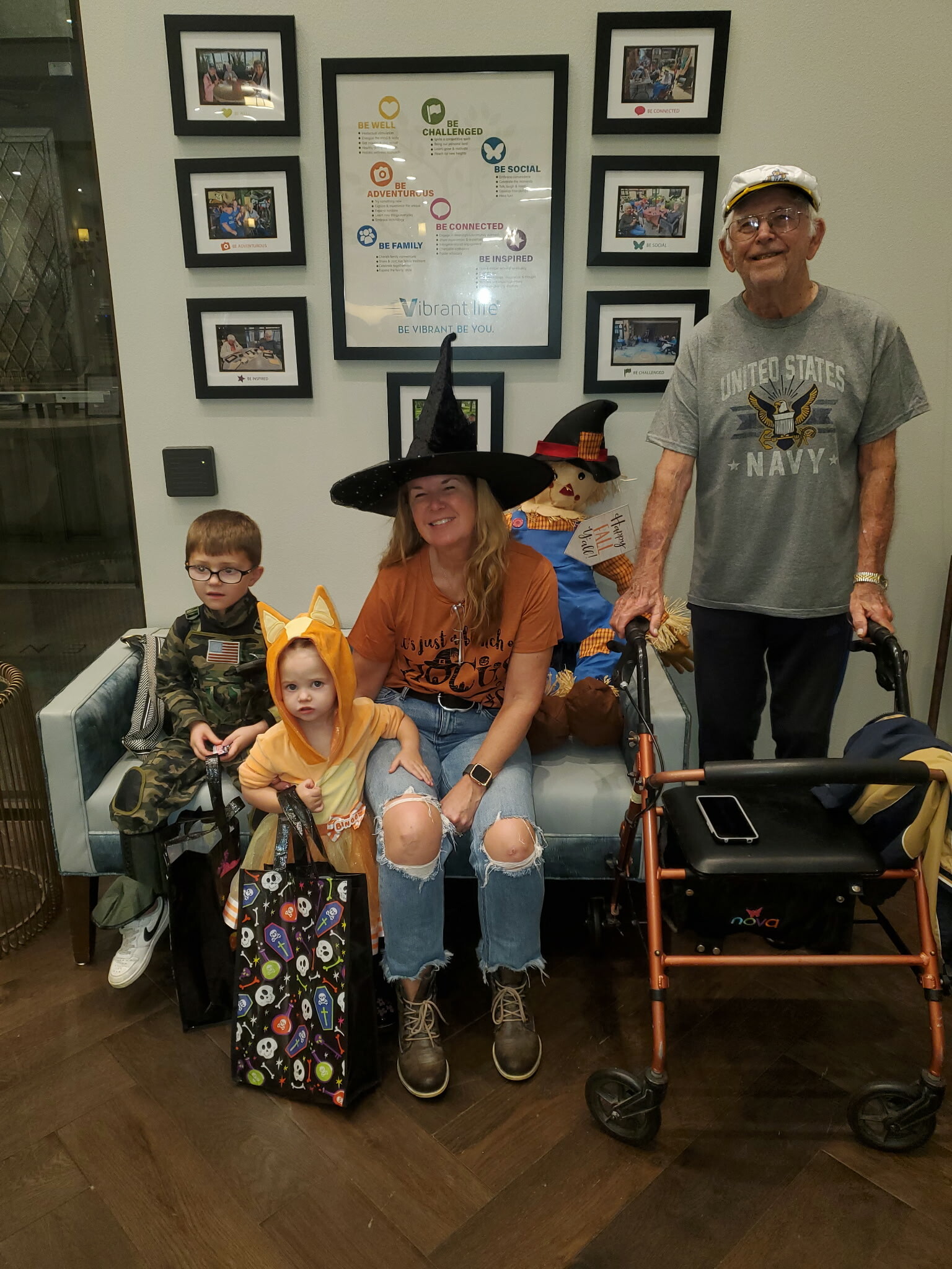 Family in Halloween costumes posing in a lobby; woman with a witch hat, kids in costumes, and an elderly man with a walker nearby.
