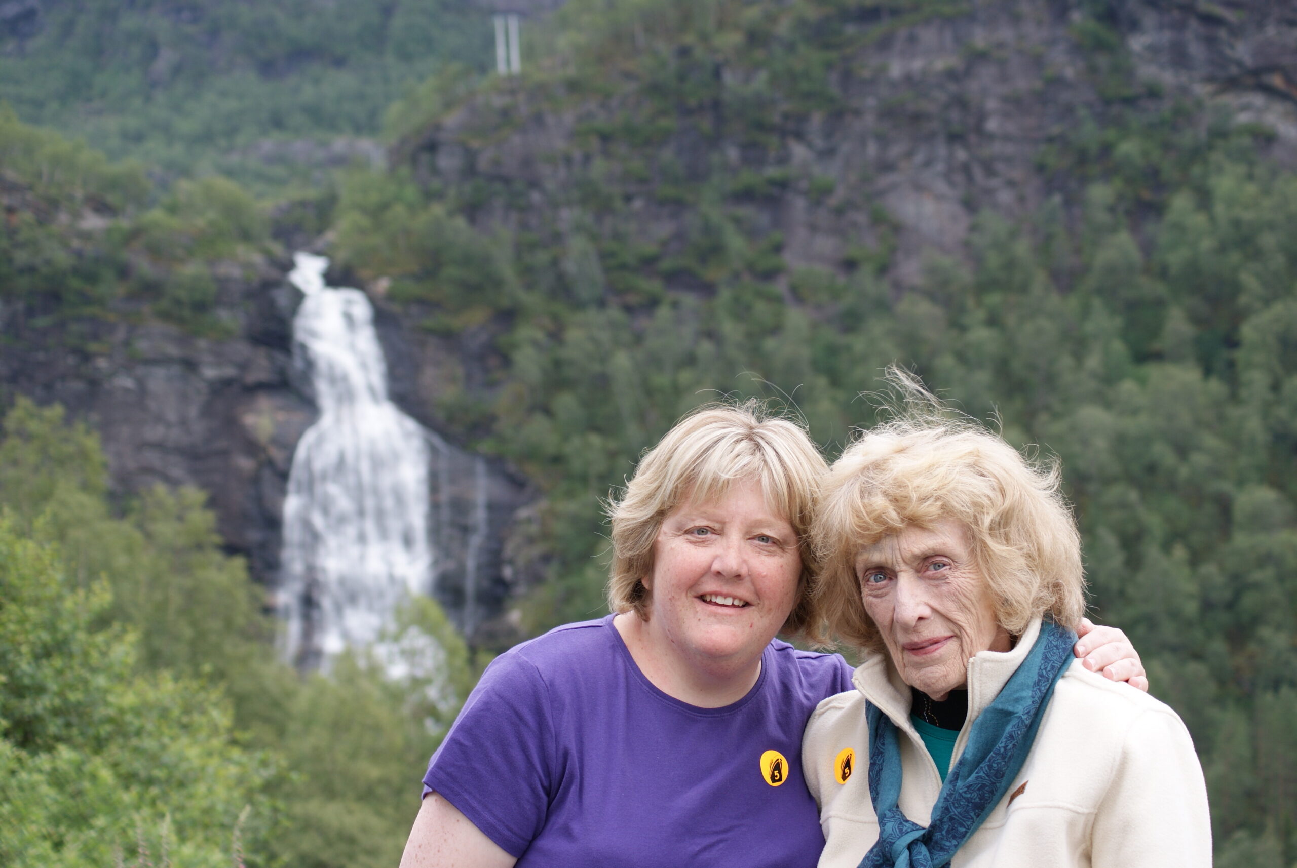 Two women smiling outdoors with a waterfall and forest behind them.