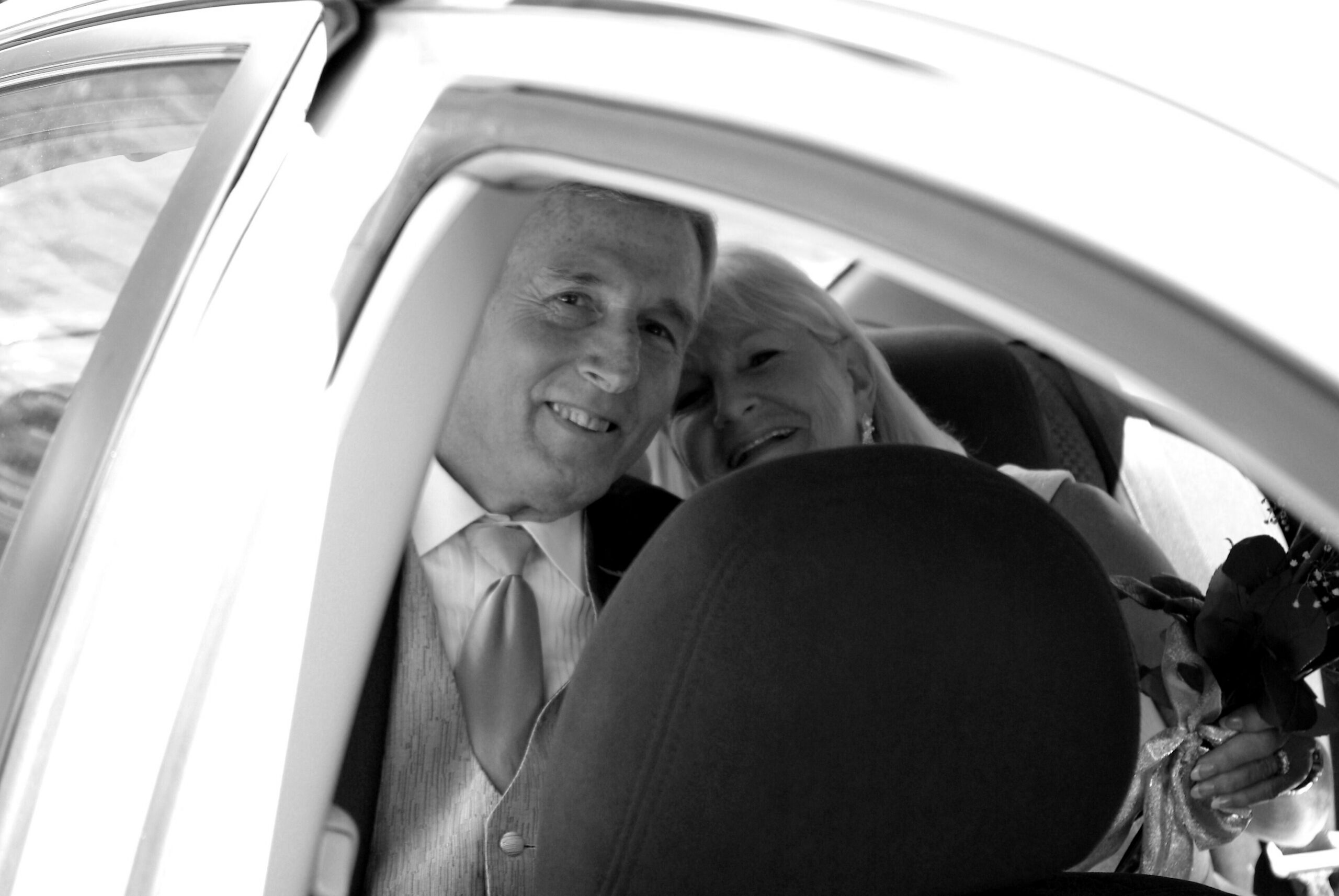 Smiling couple leaning from a car window, man in a suit and woman beside him, posing for a photo.