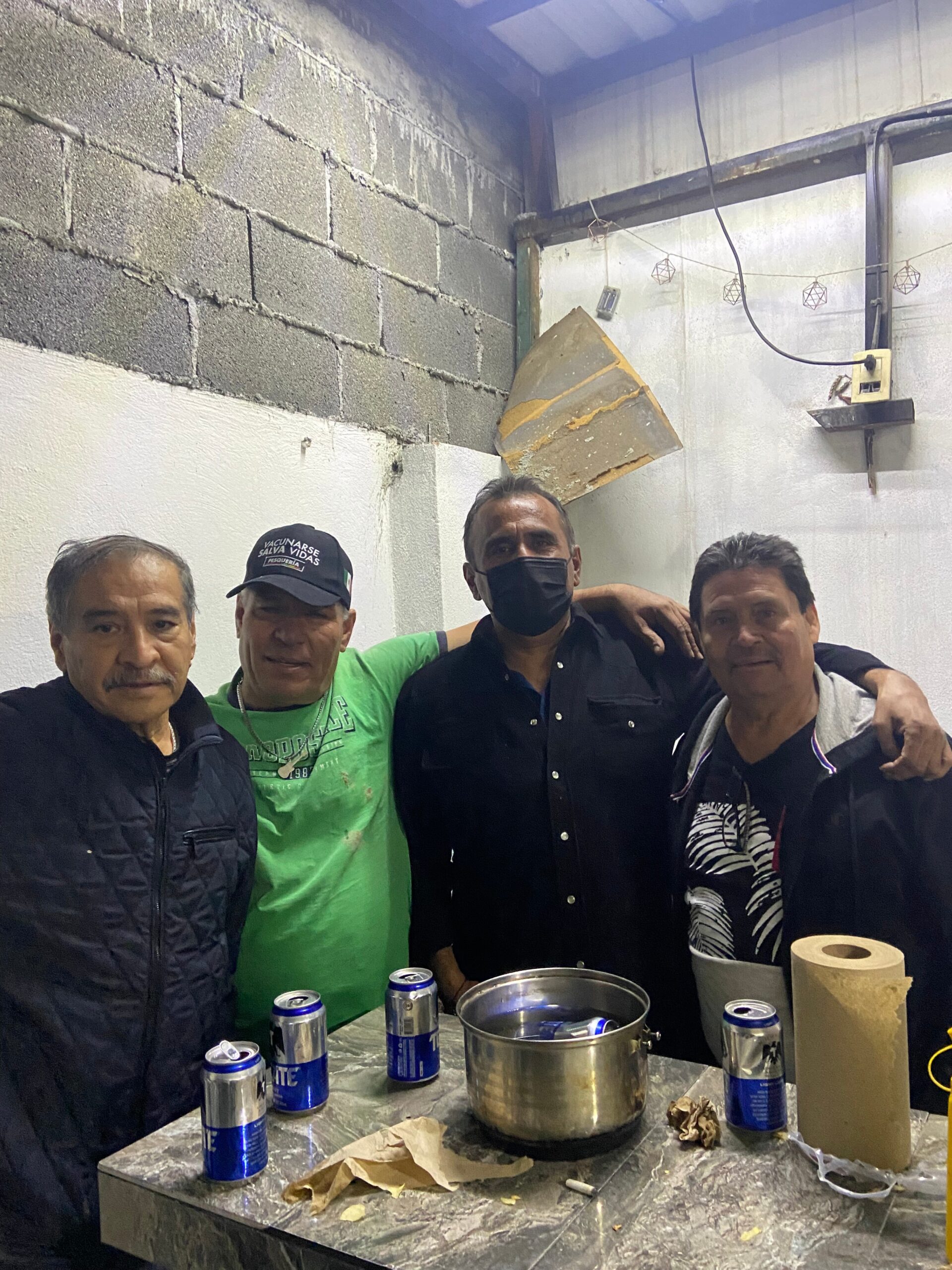 Four men pose in a dim workshop with a concrete block wall, cluttered metal table, and metal pot in front.