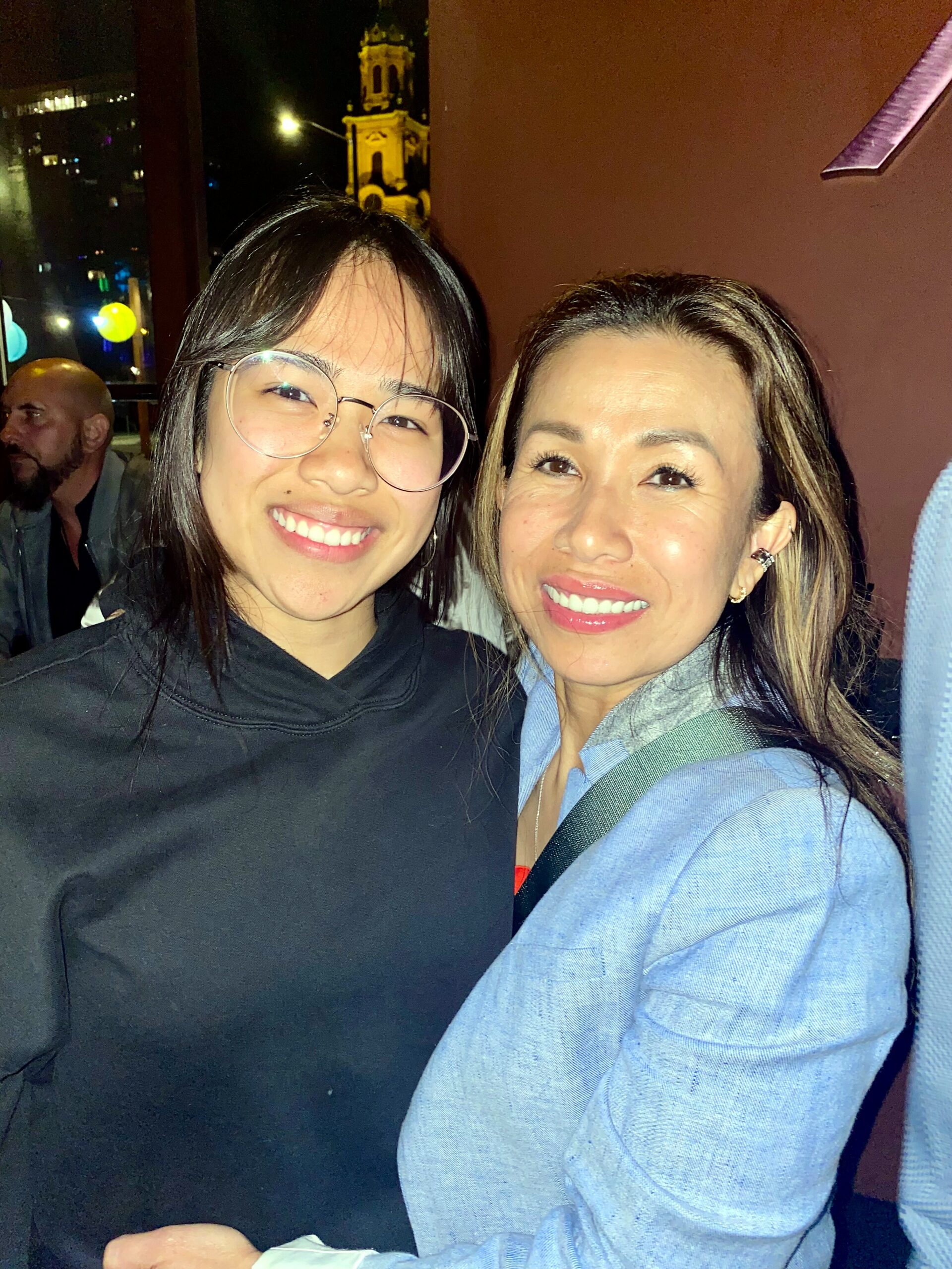 Two women smiling for a selfie at night, one wearing round glasses and a black top, the other in a light blue jacket.