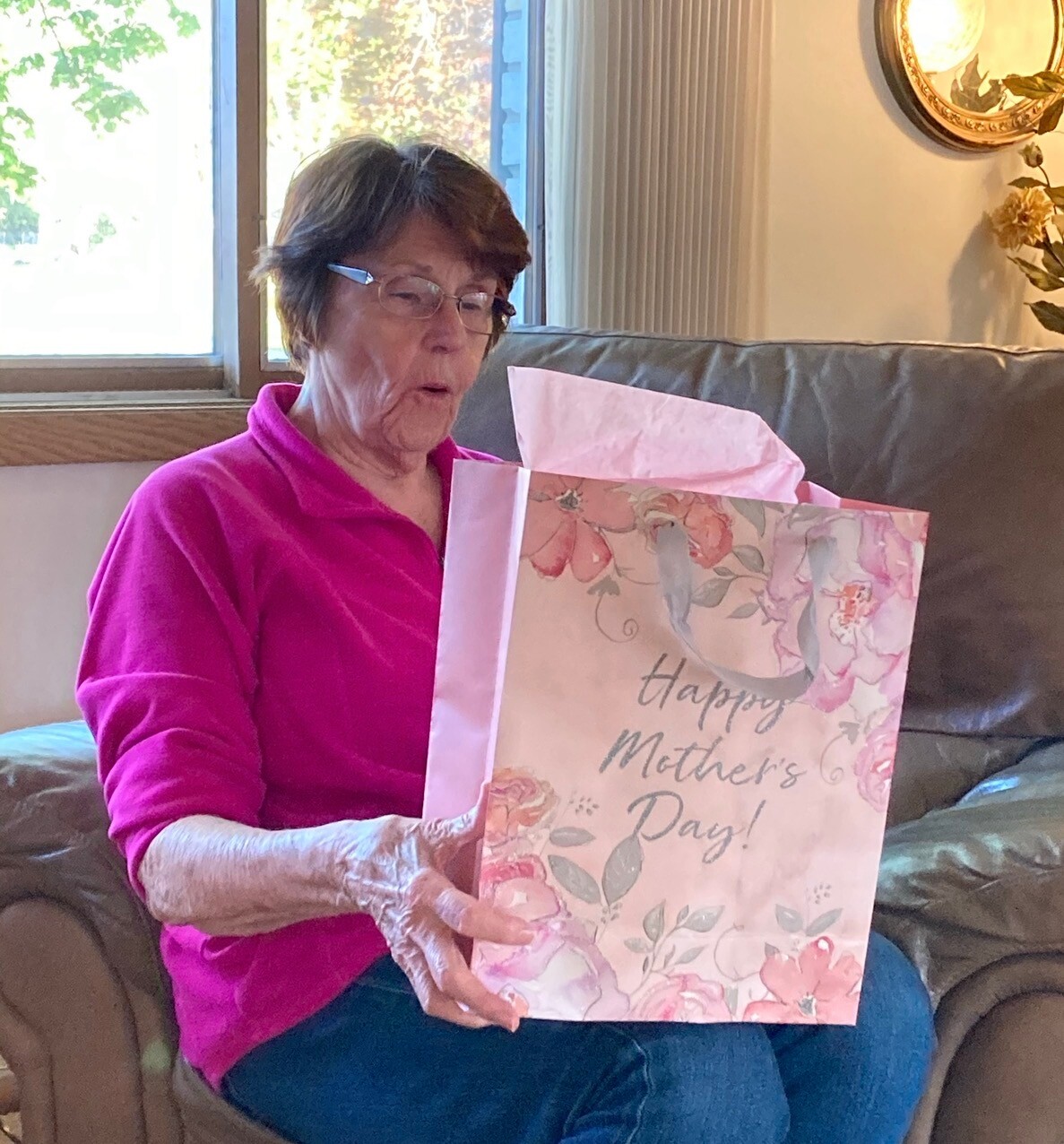 Older woman in a bright pink top sits on a couch, looking at a pink floral gift bag that says 'Happy Mother's Day'.