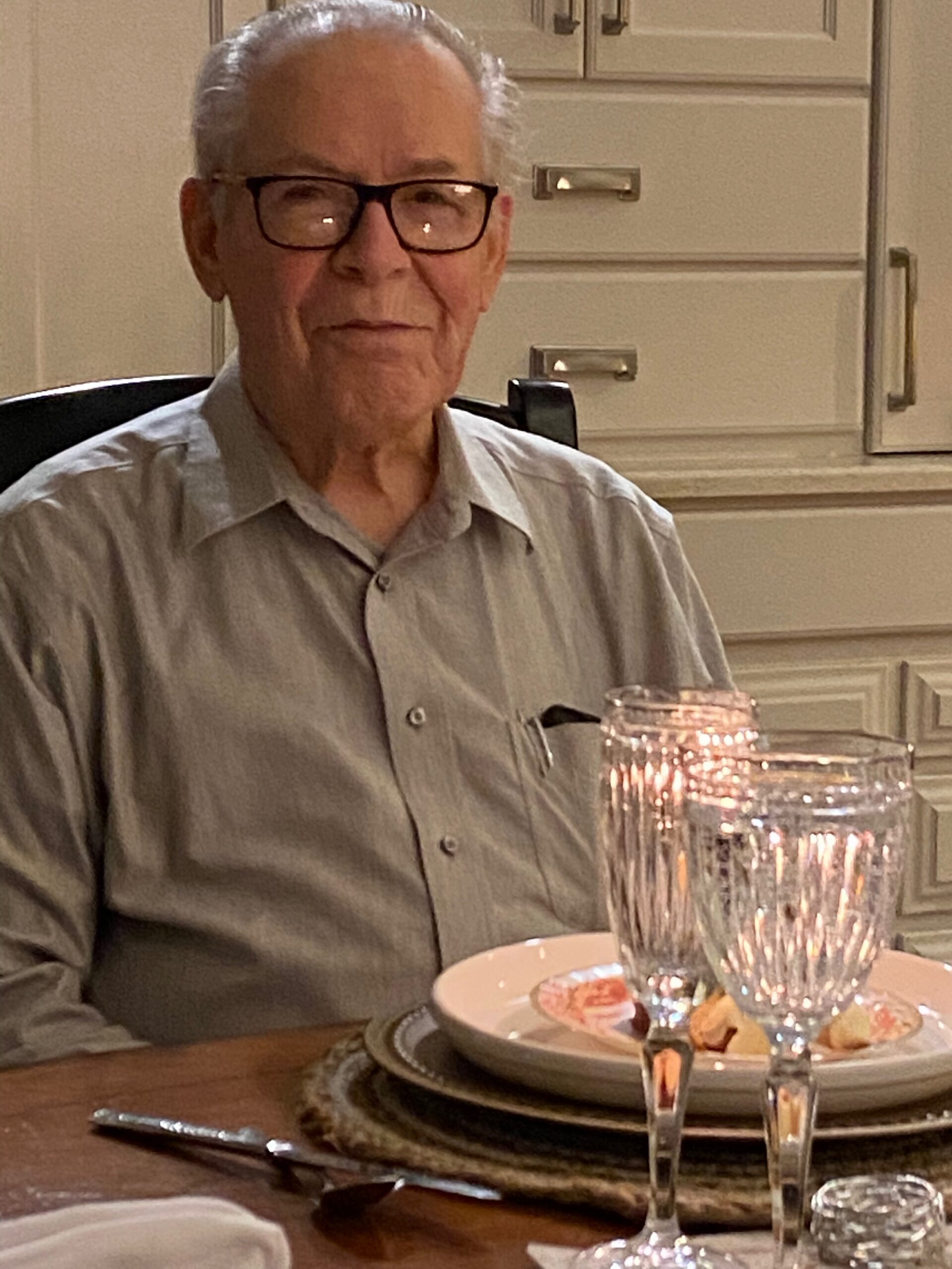 Older man with gray hair and glasses sits at a dining table, with a plate, utensils, and crystal candle holders in view.