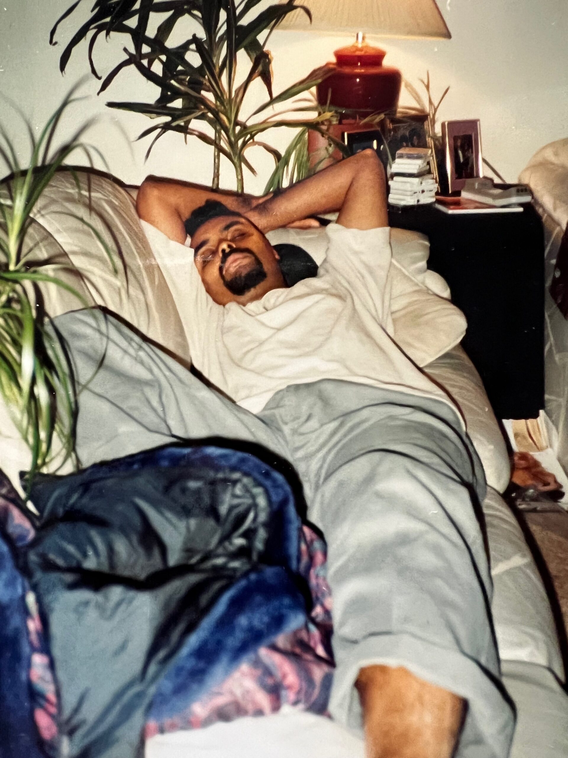 Man asleep on a beige sofa with arms behind his head, surrounded by a tall plant, lamp, and a stack of books on the side table.