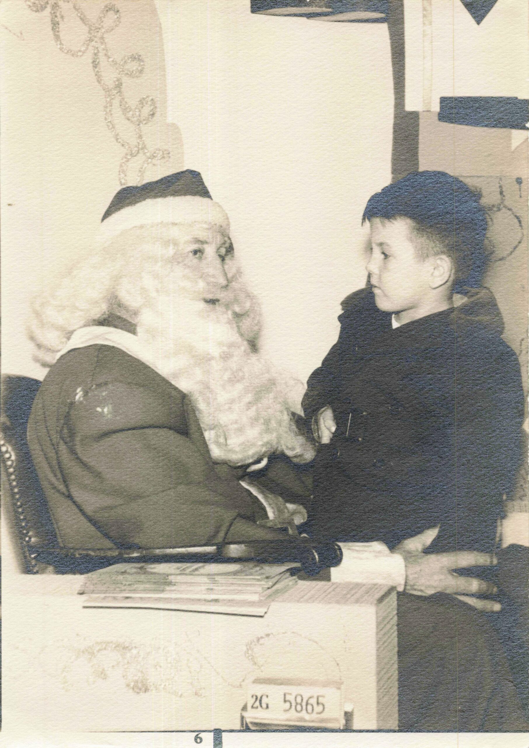 Santa Claus with a white beard talking to a young boy indoors, papers on a desk nearby.