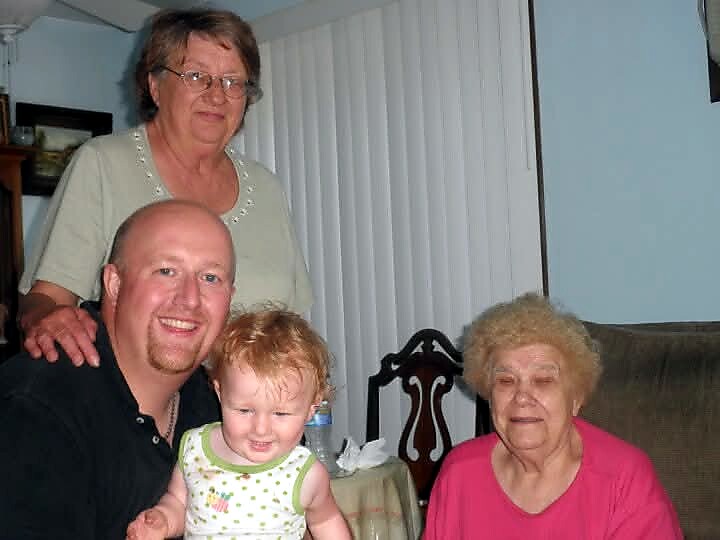 Group photo: a man holding a smiling baby girl, with an older woman standing behind them and a grandmother seated nearby in a living room.