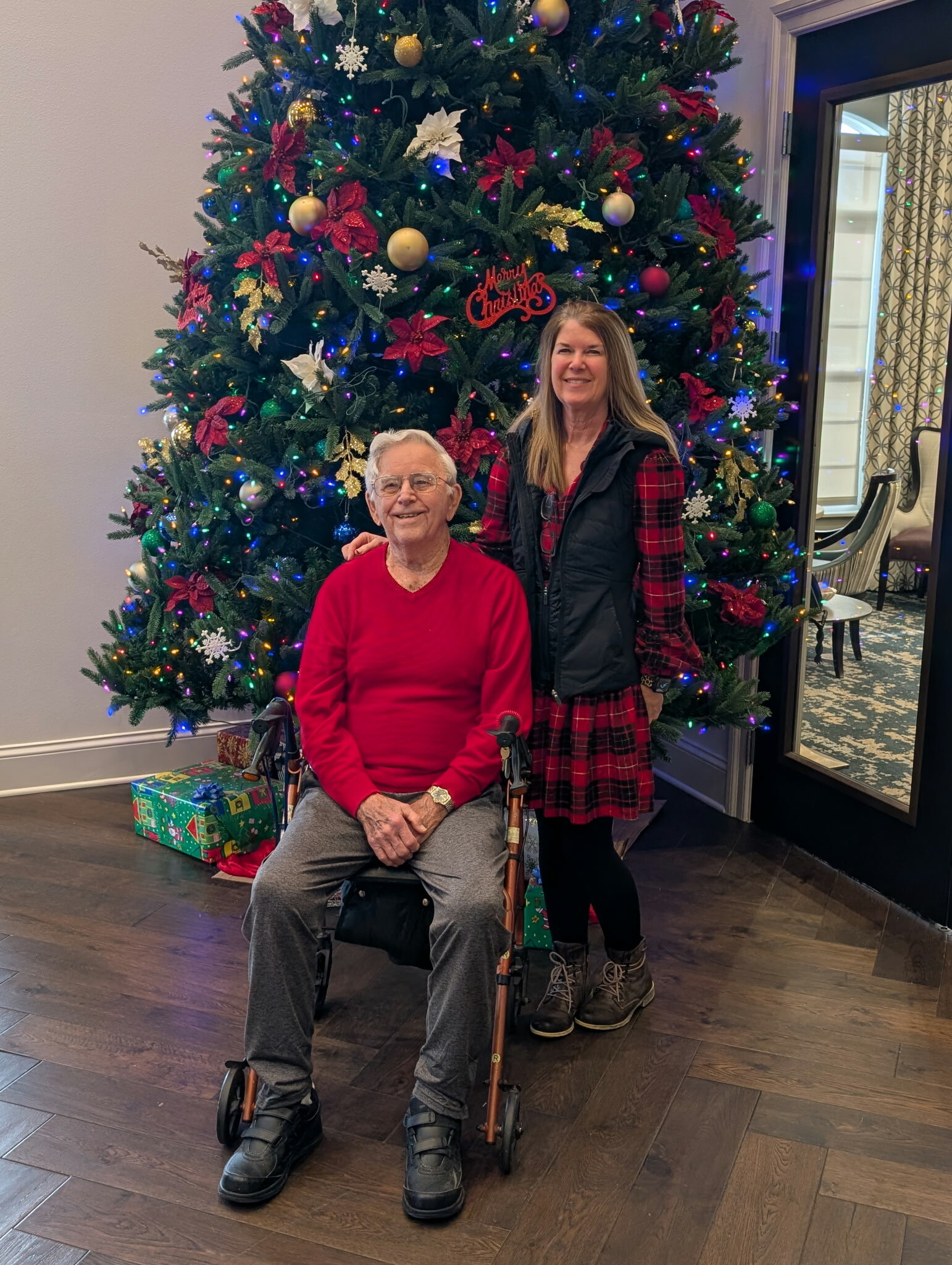 Elderly man in a red sweater sits in a wheelchair beside a decorated Christmas tree; a smiling woman in a plaid dress stands behind him.
