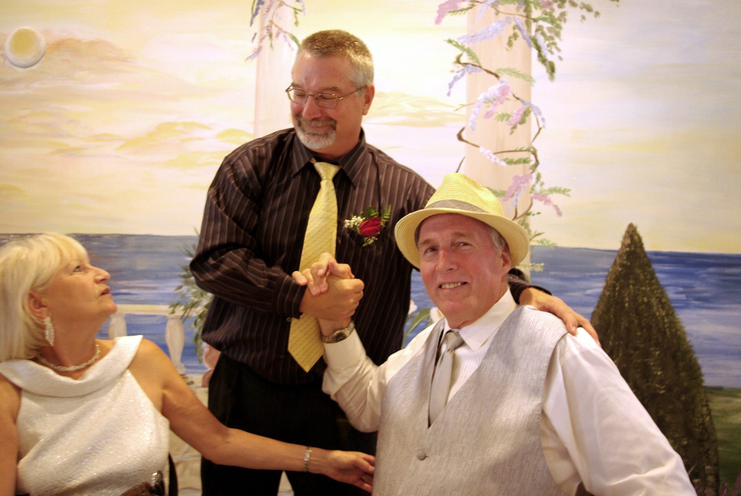 Three adults dancing together at a festive celebration, smiling against a tropical backdrop with palm trees and a painted sea.