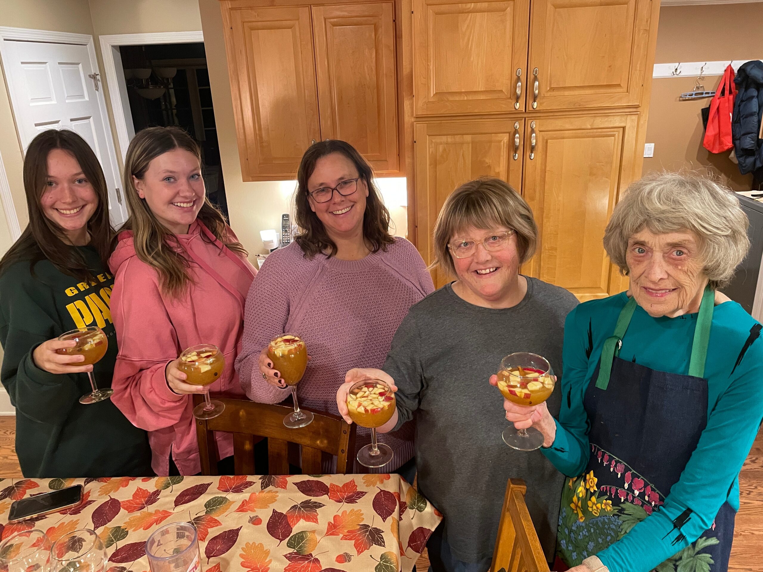 Five women standing by a kitchen island, smiling, each holding a stemmed glass with an orange fruit cocktail and ice. Autumn leaf-patterned tablecloth in foreground and wooden cabinets in the background.
