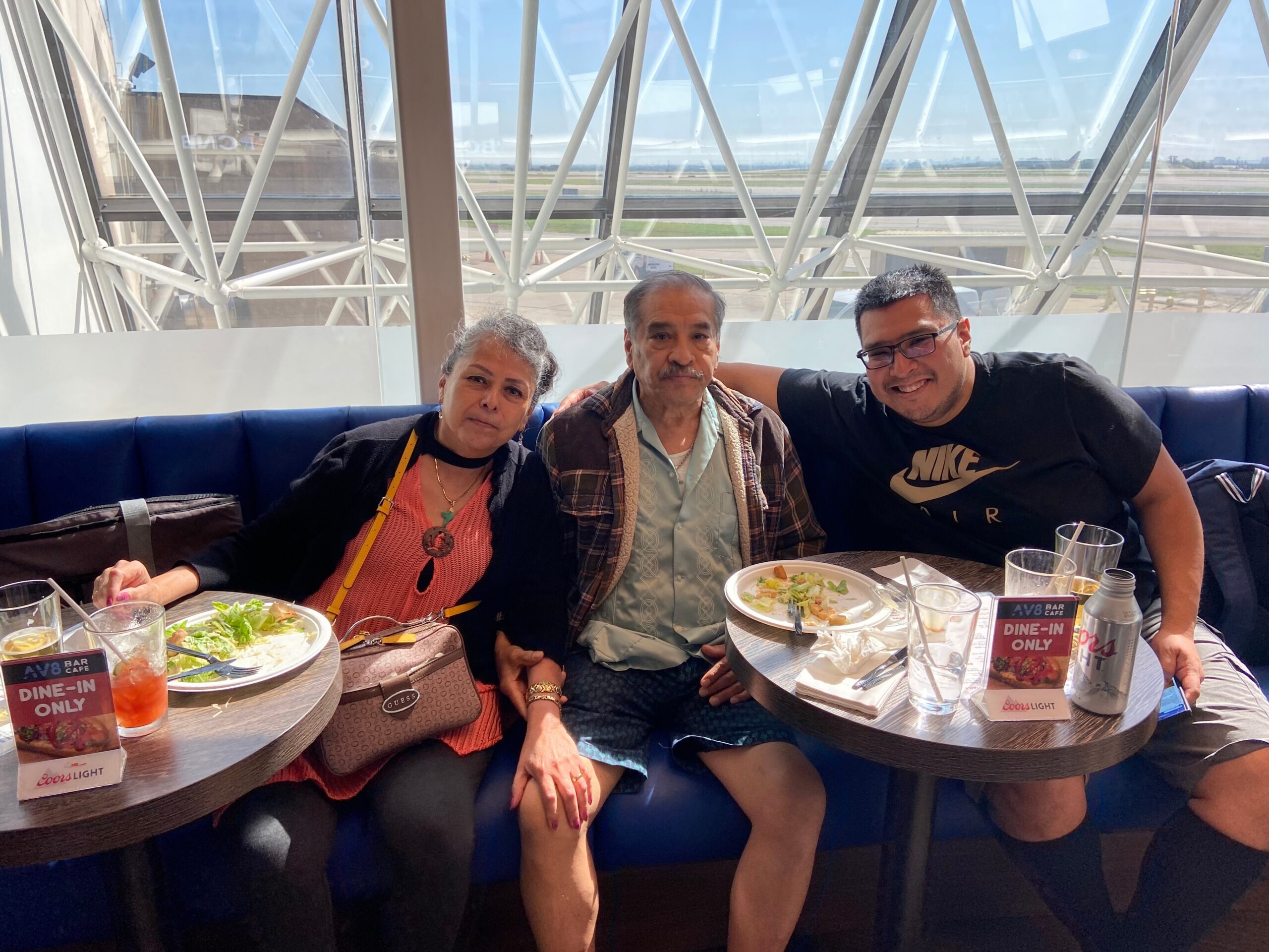 Three people sit closely together at a round table, enjoying salads in a bright, modern dining area with a blue cushioned bench behind them and a glass-and-metal architectural background behind.
