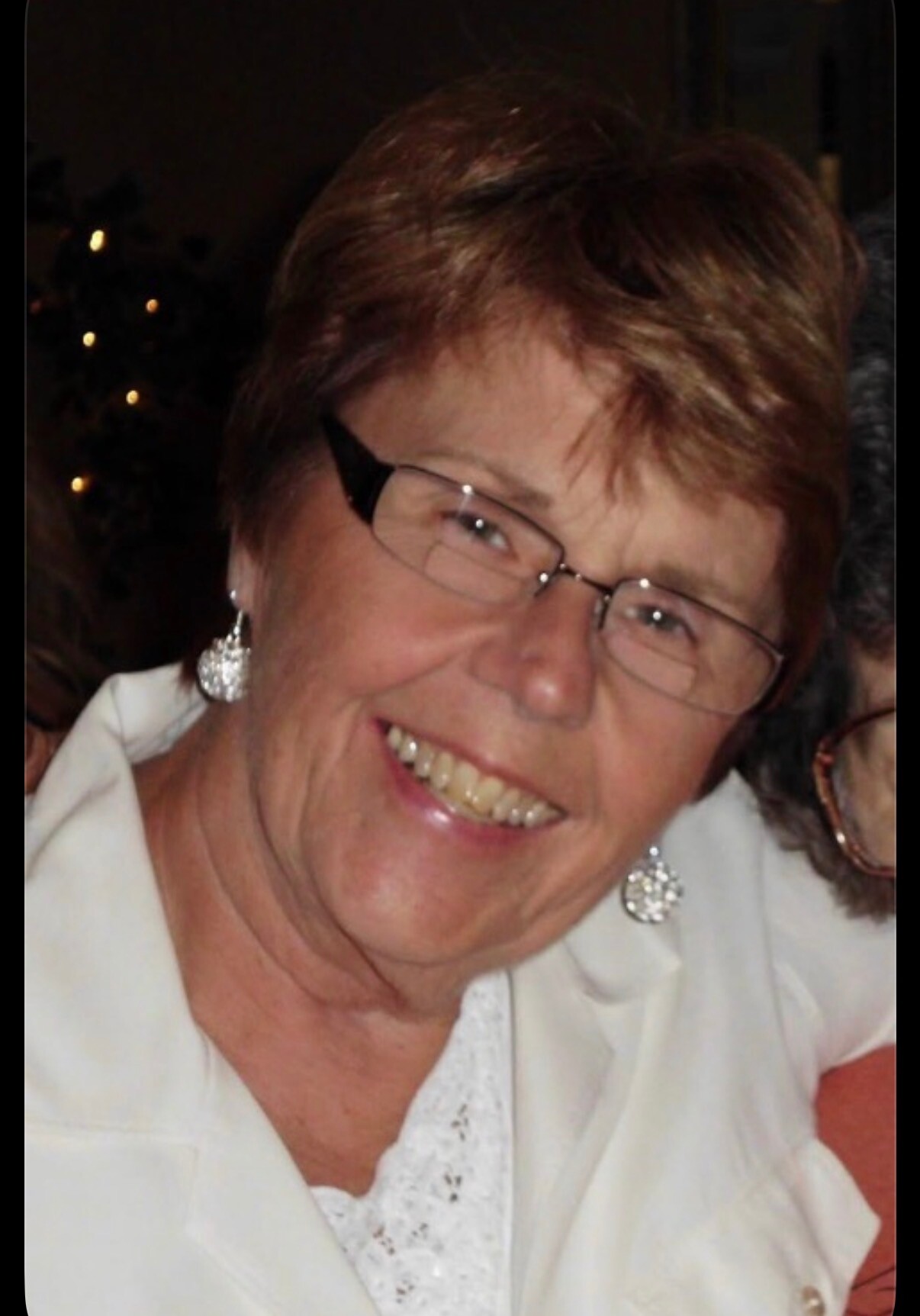Smiling woman with short brown hair, glasses, and dangling earrings in a white blouse.