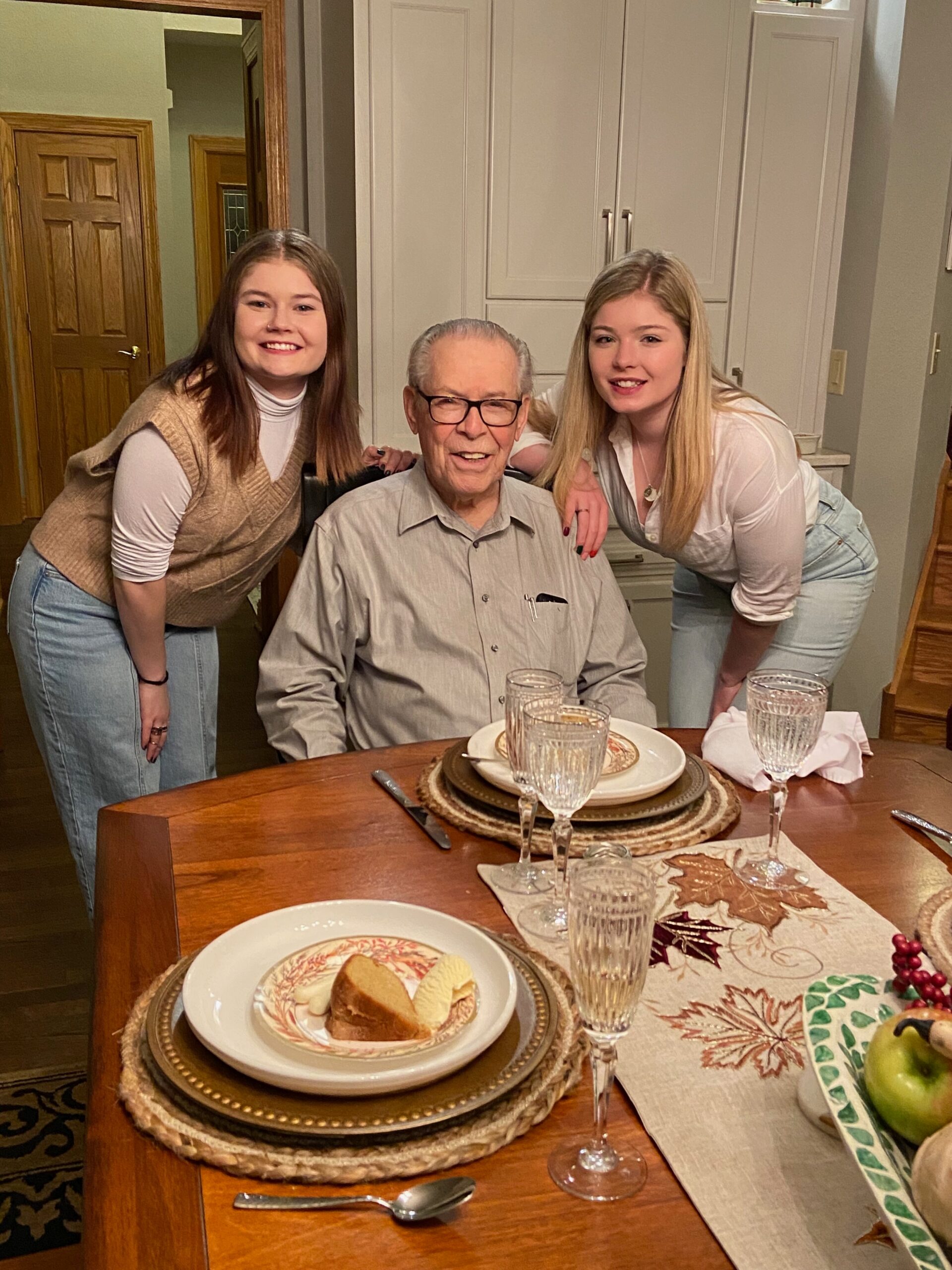 Smiling elderly man seated at a dining table, flanked by two young women leaning in, in a home kitchen setting.