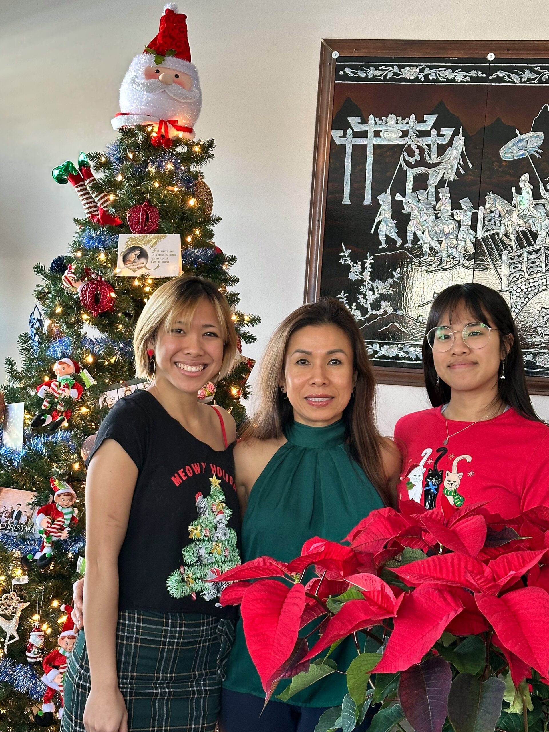 Three women smiling in front of a decorated Christmas tree indoors.