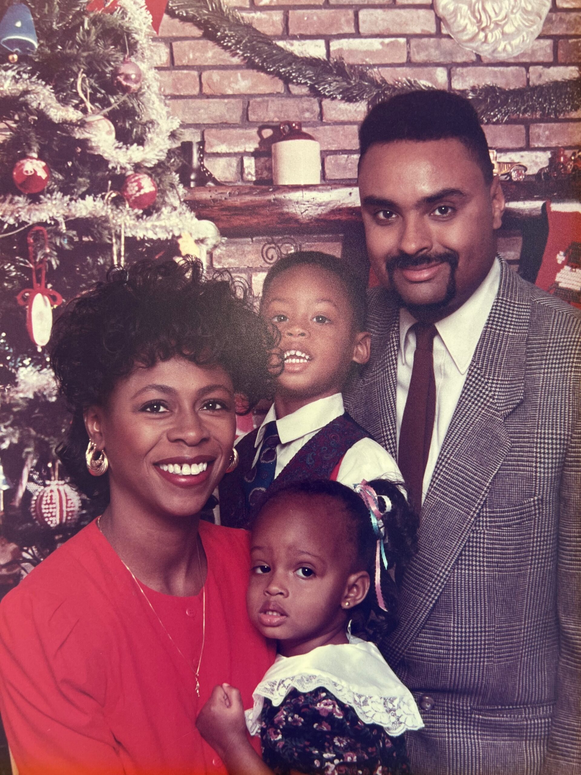 Family portrait of an African American mother, father, and two children smiling beside a decorated Christmas tree and brick fireplace; festive holiday scene.
