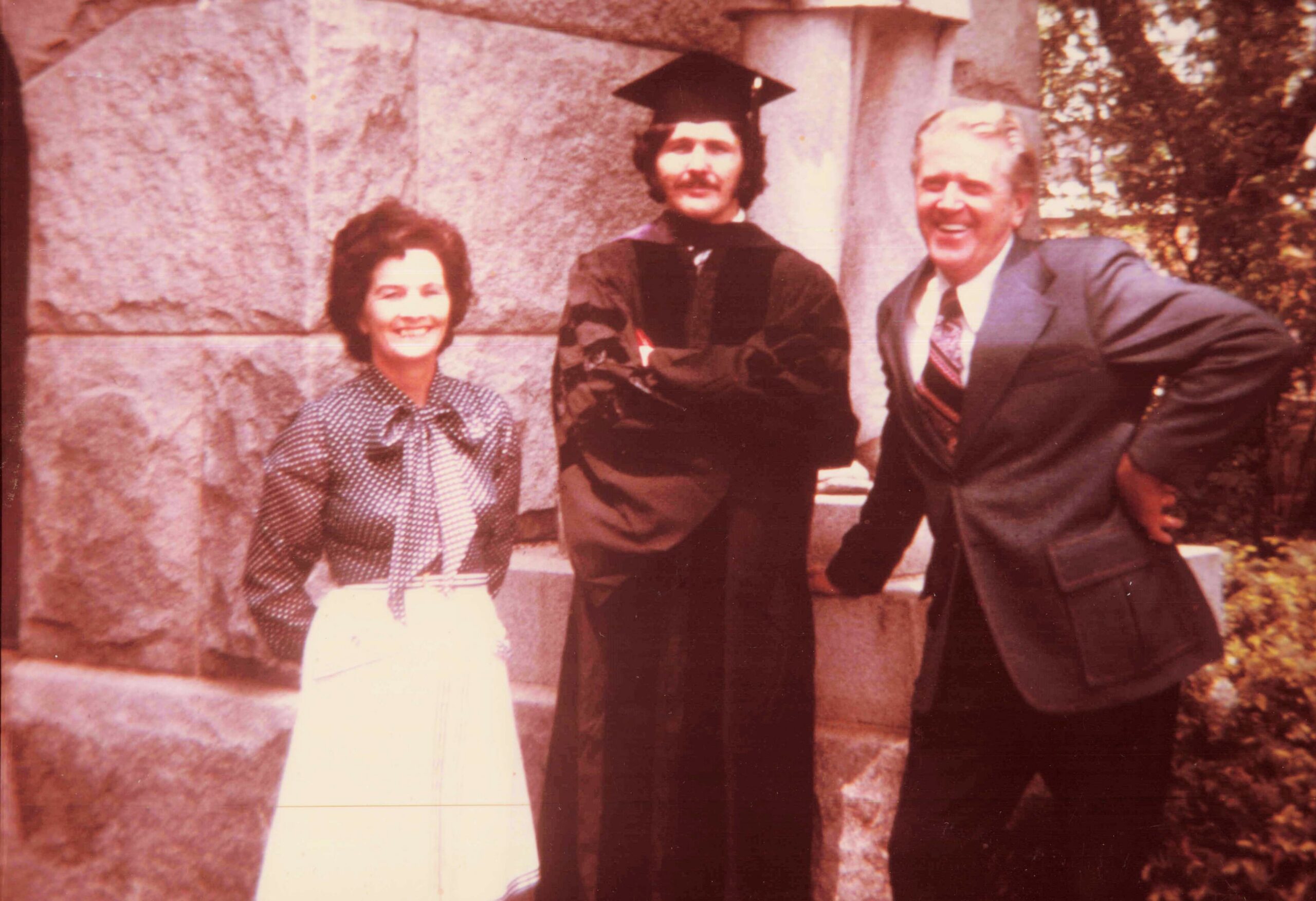 Woman in a polka-dot blouse and white skirt smiles beside a man in a graduation gown and cap, with another suited man on a stone steps backdrop outdoors.