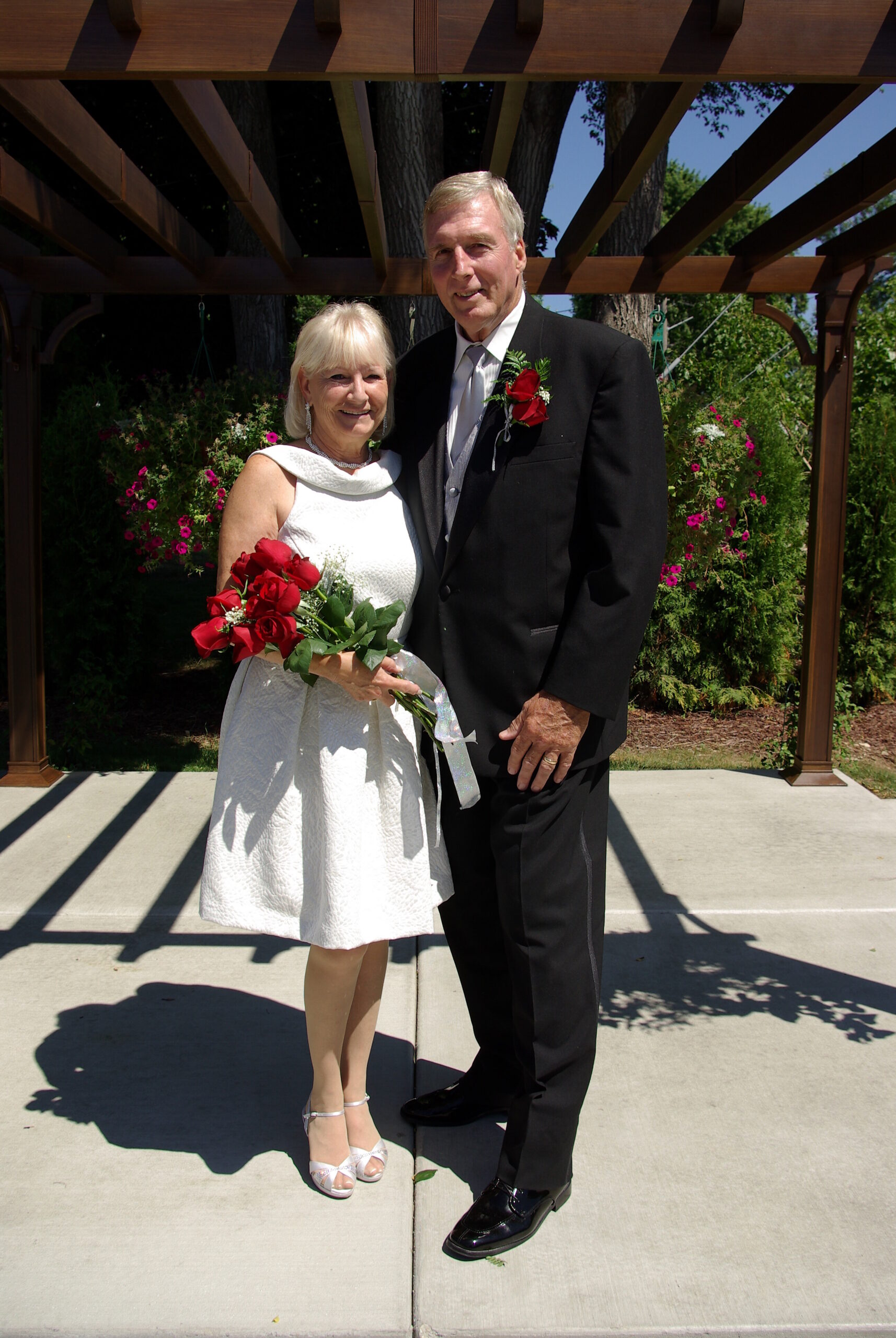 Elderly couple posing outdoors in formal attire; woman in a white dress holding a red rose bouquet, man in a black suit with a boutonniere under a wooden pergola.