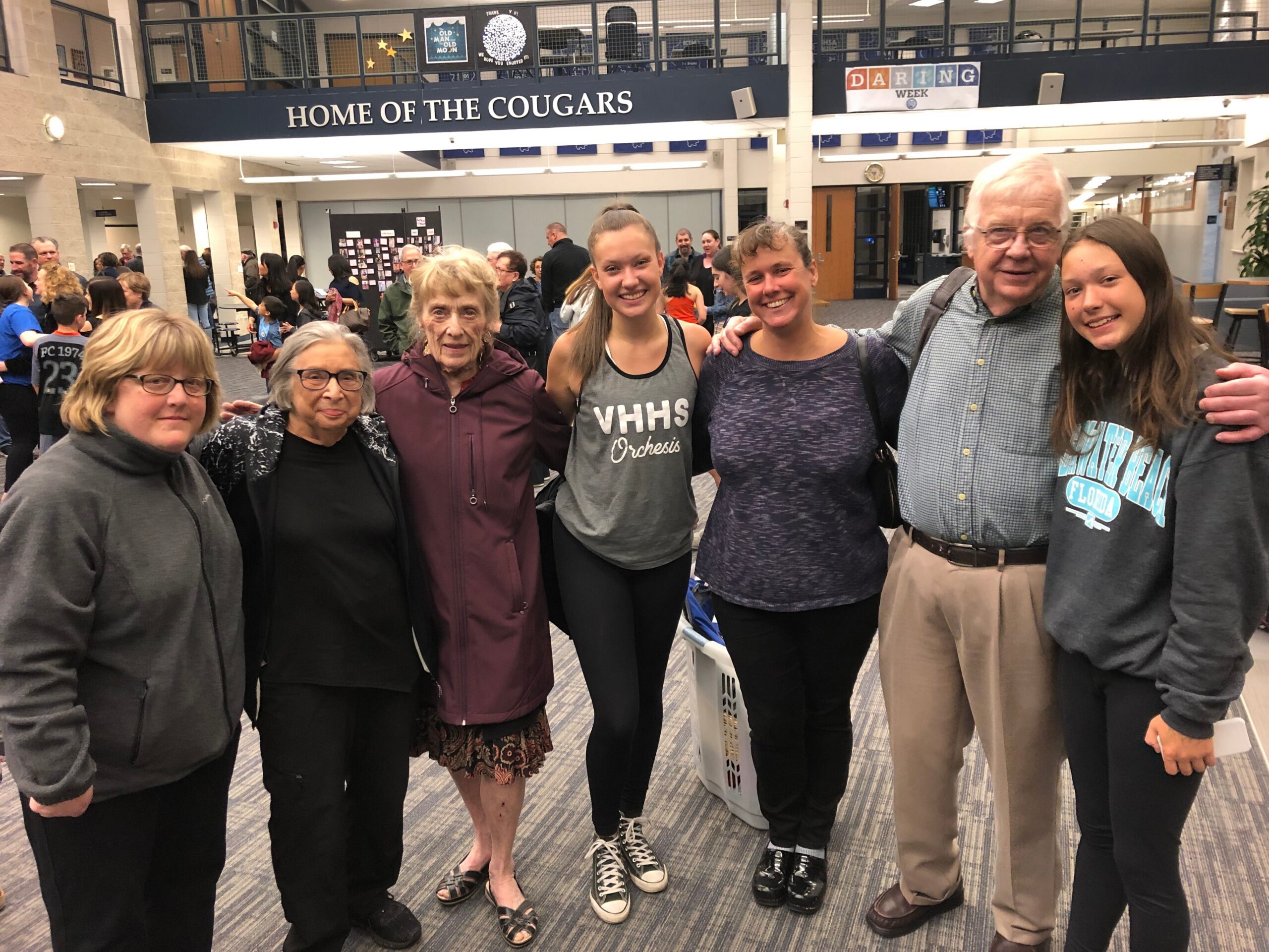 Group of people smiling for a photo in a school lobby, with 'HOME OF THE COUGARS' sign overhead and students and adults gathered in the background.