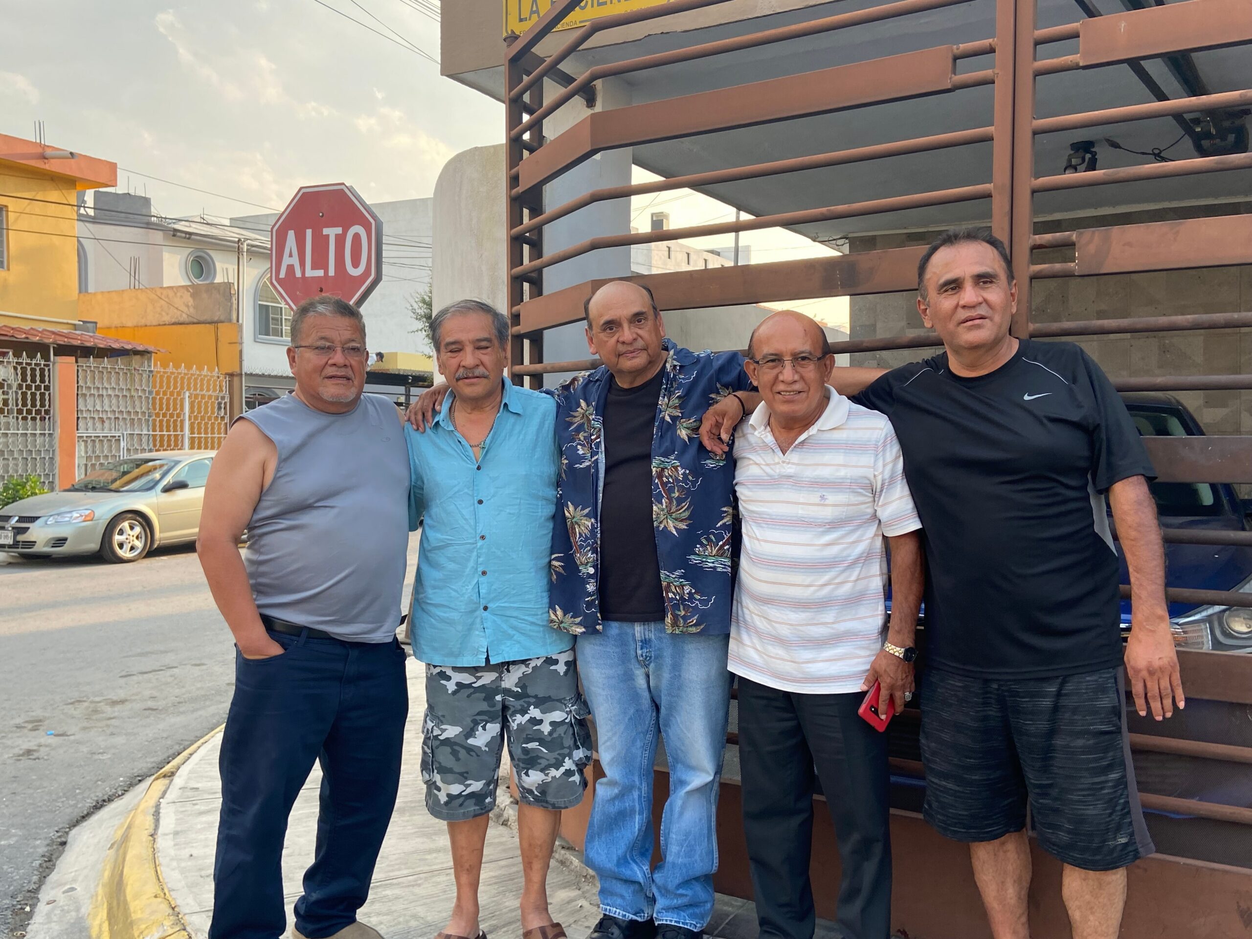 Five men stand together with arms around each other on a street, smiling at the camera; casual summer clothing, near a brown metal railing and a stop sign in the background.