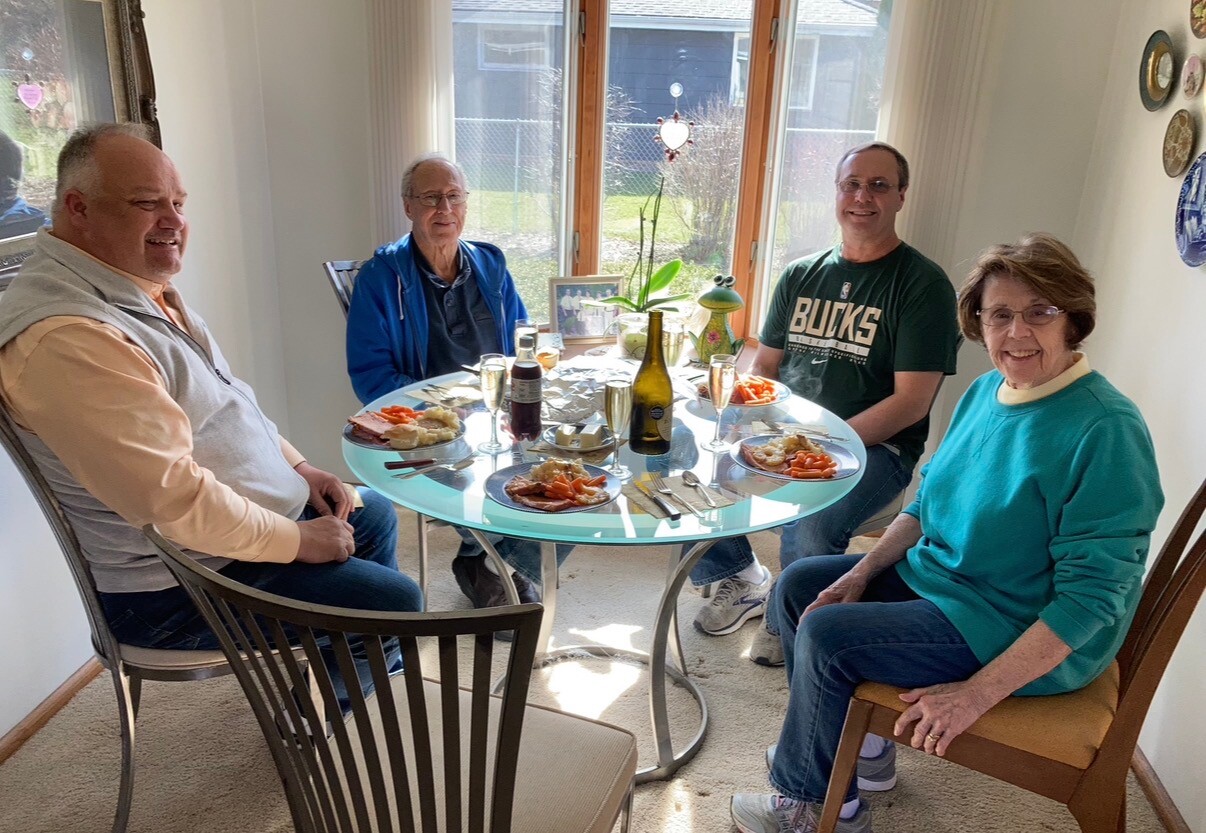Four adults sit around a glass round table with plates of food, drinks, and a bottle in a sunlit dining area.