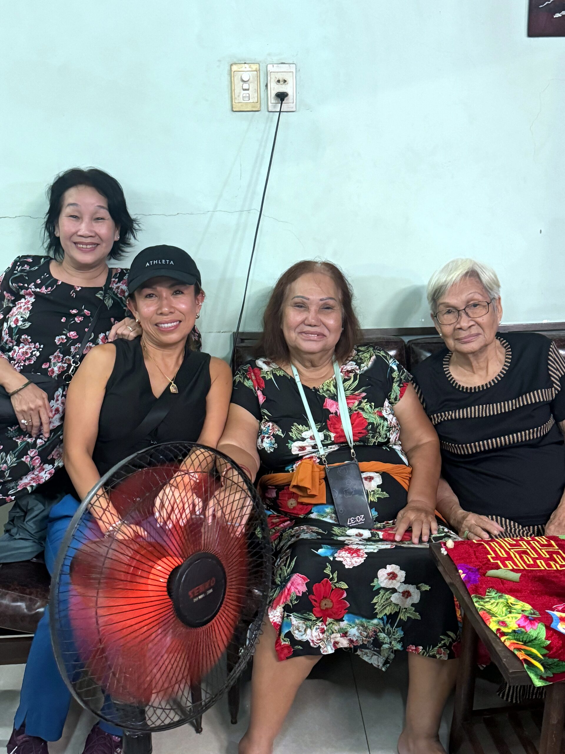 Four women posing for a photo indoors, colorful floral dresses, with a red electric fan in the foreground and a light blue wall behind them.