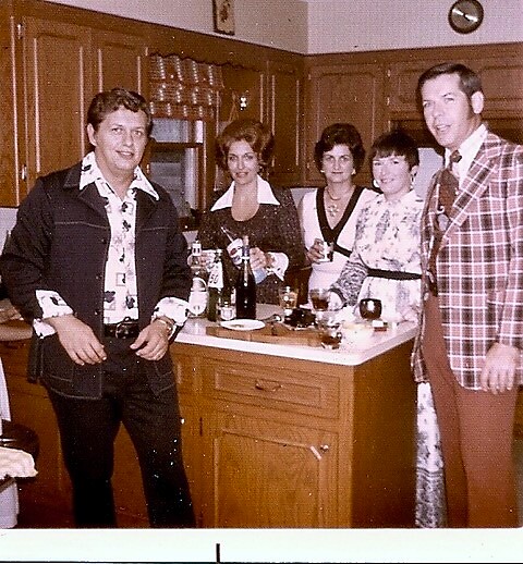 Five adults in a retro 1970s kitchen posing around a counter with drinks on the island and a relaxed vibe.