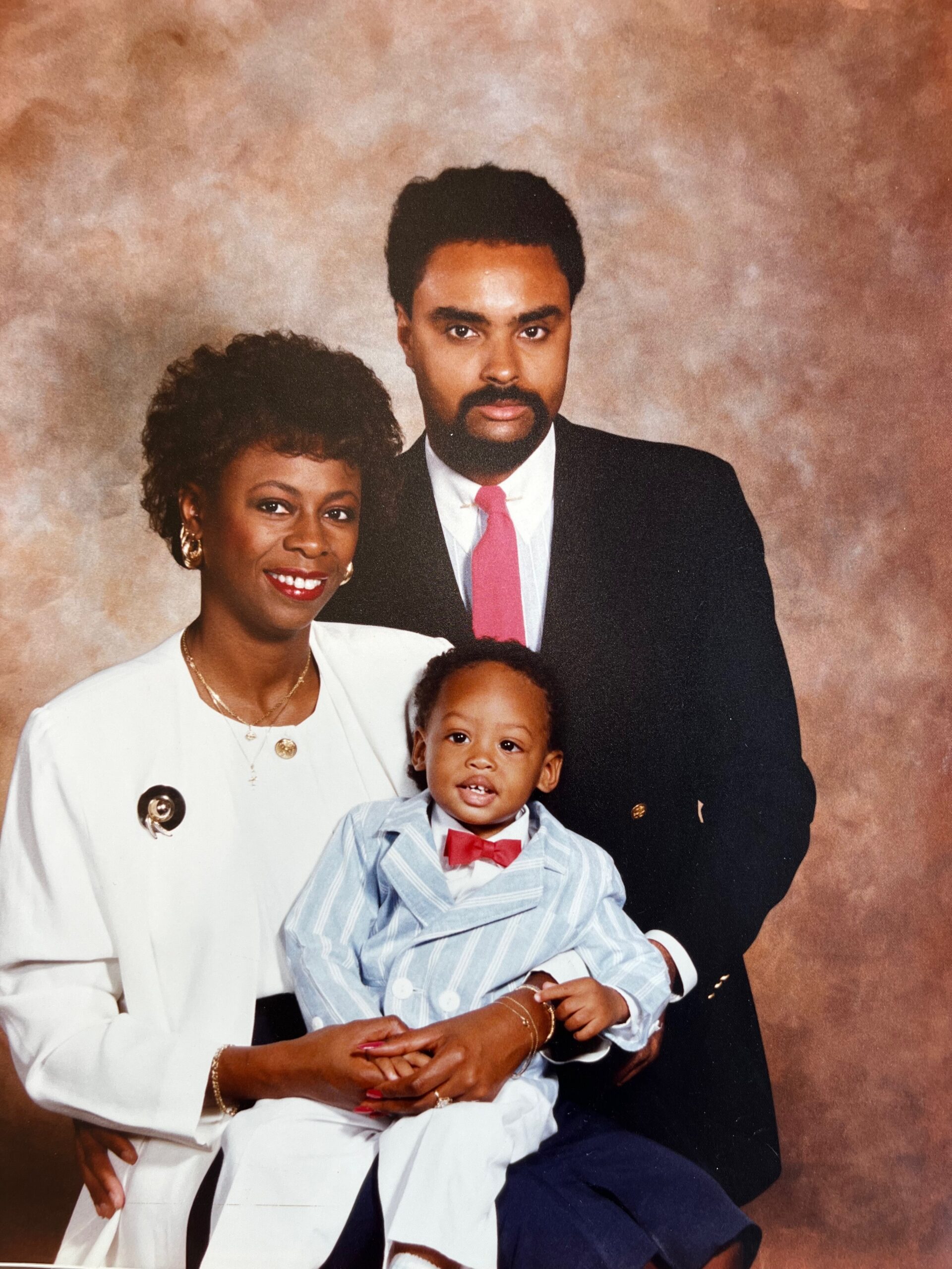 Family portrait of a mom, dad and toddler posing together in a studio backdrop, dressed in formal attire.