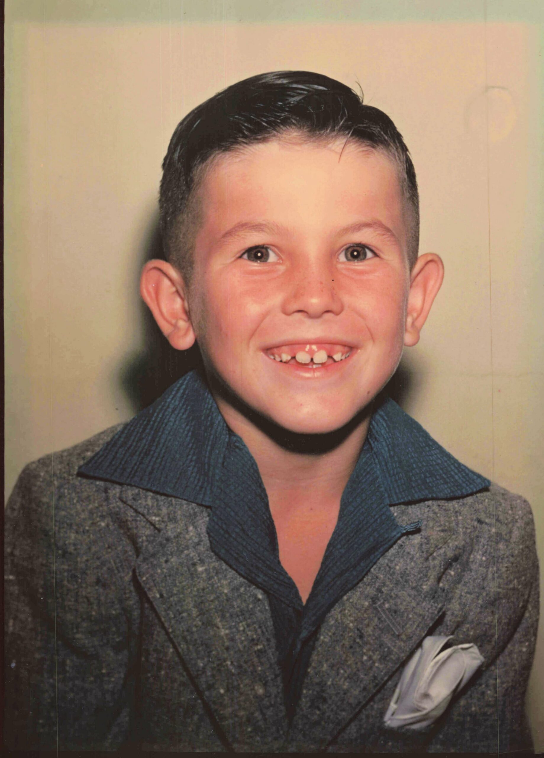 Smiling boy in a gray blazer with a blue collar, looking at the camera and showing teeth.