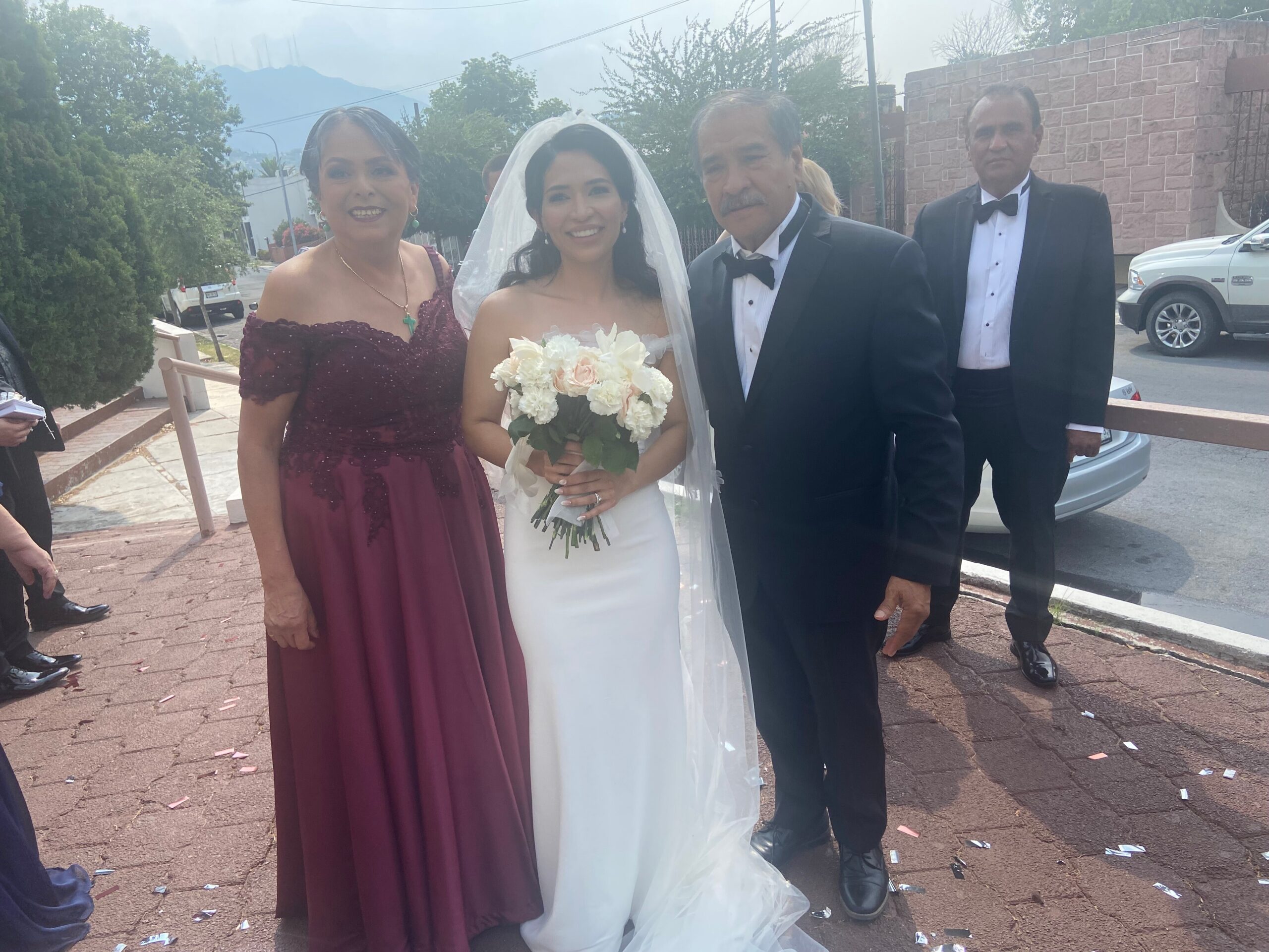 Bride in white gown and veil holding a bouquet of pale pink and white flowers, flanked by a woman in a burgundy formal dress and a man in a black tuxedo outdoors, with confetti on the ground after a ceremony.