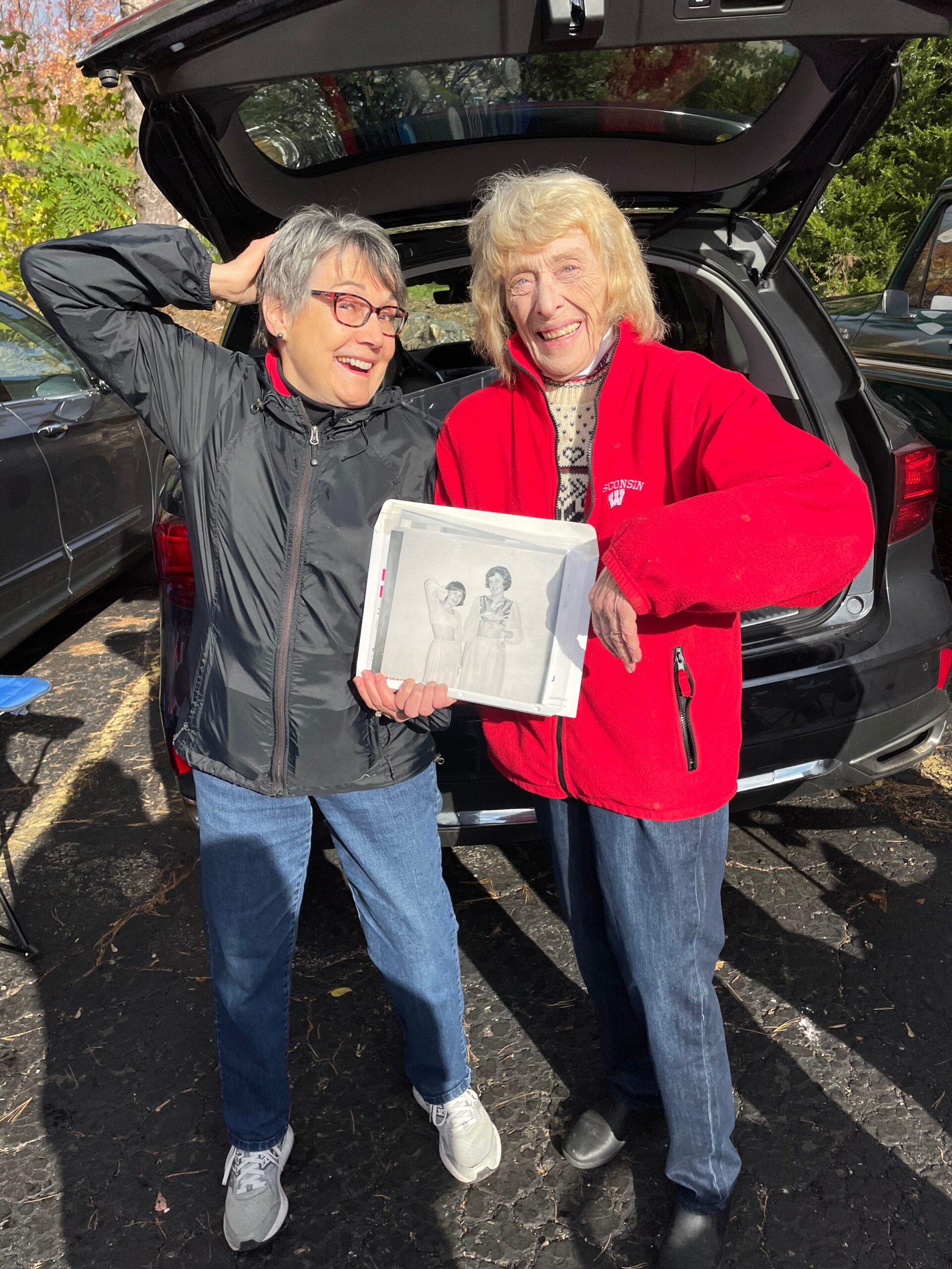 Two women smiling in a parking lot by an open SUV trunk; one in a black jacket holding a photo album showing fashion images.