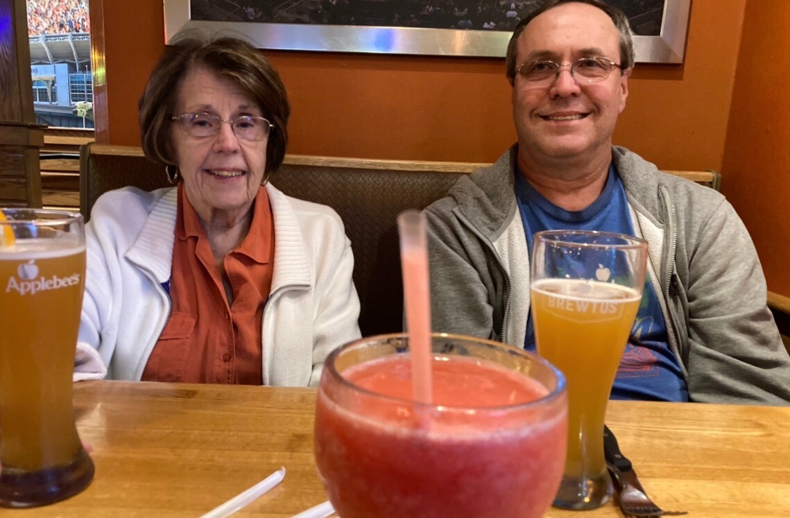 Smiling elderly woman and man sit at a booth with two beer glasses and a pink frozen drink in front of them at a restaurant.