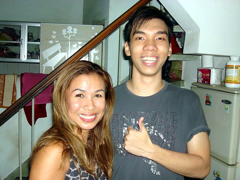 Two friends pose for a photo indoors; the man gives a thumbs-up and both smile at the camera in front of a staircase and fridge.