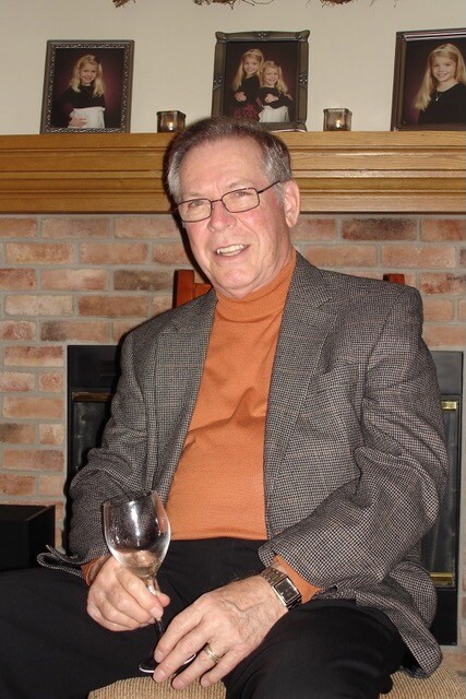 Older man in a gray blazer and orange turtleneck sits by a brick fireplace, holding a wine glass and smiling.